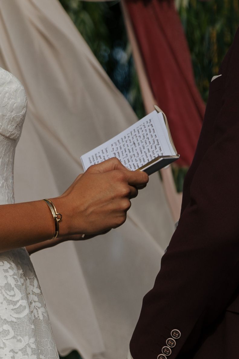 Close-up of hands writing wedding vows in a notebook against a white dress and burgundy background.