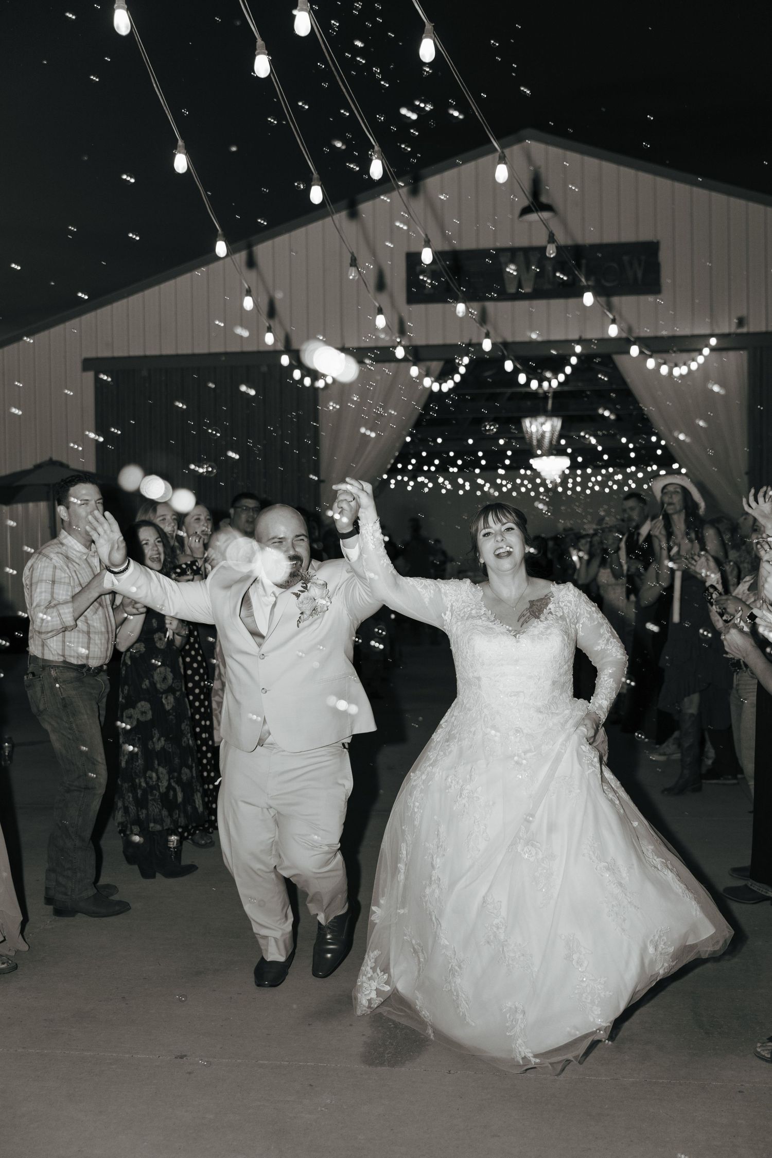 Black and white photo of a bride and groom dancing joyfully under string lights at their wedding reception.