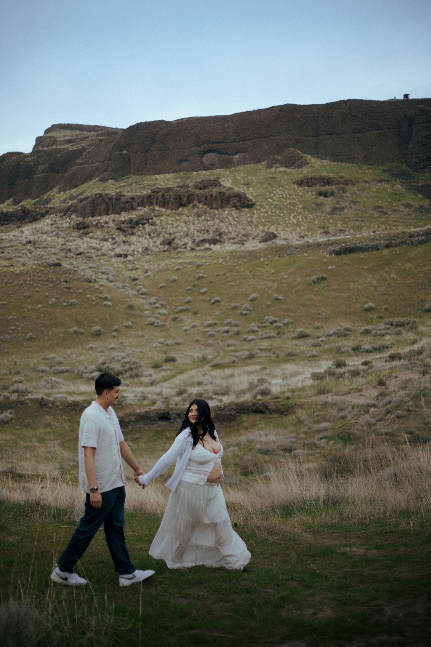 Couple playfully runs through a grassy hillside landscape with dramatic cliffs in the background.