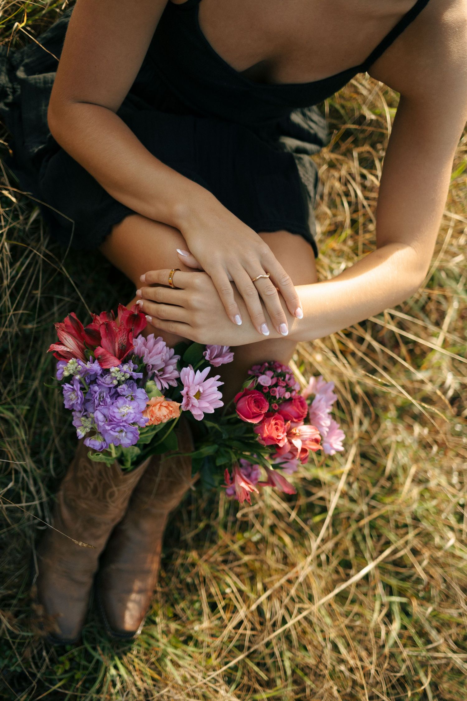 Hands holding colorful flowers against a rustic background with brown boots and straw visible in sunlit setting.