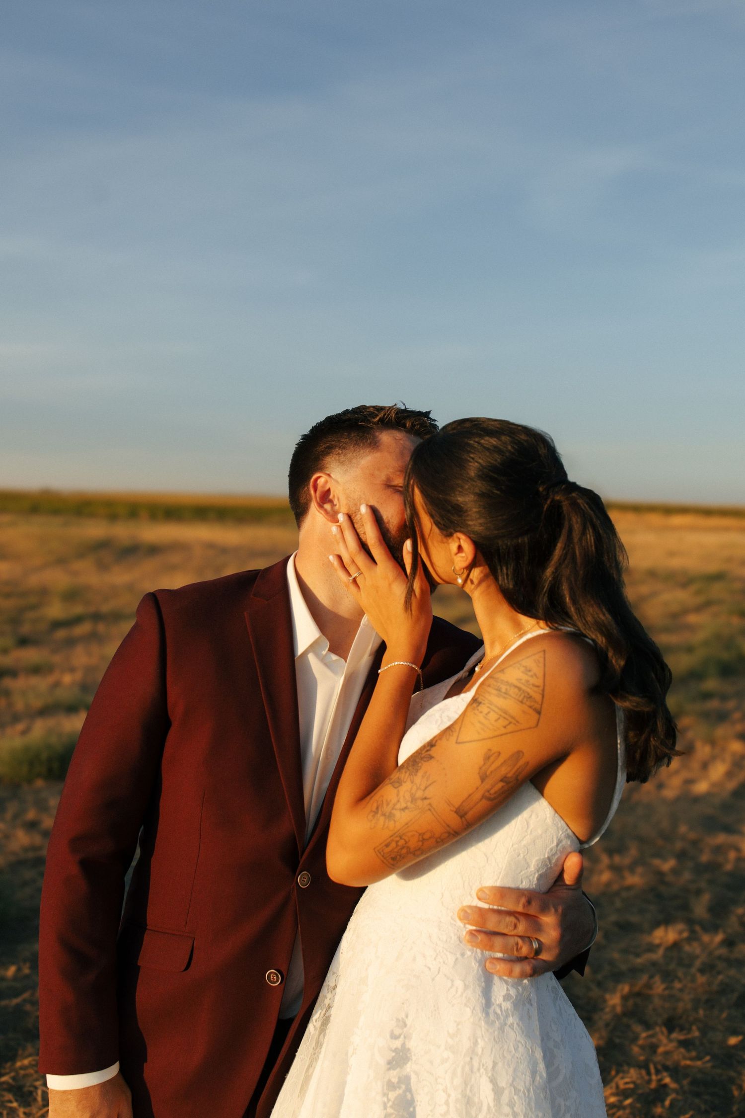A romantic couple in wedding attire share intimate kisses at sunset in an open field.