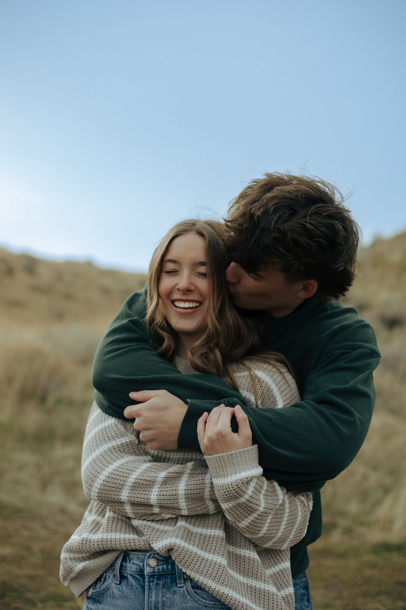 A couple laughs and embraces while wearing casual sweaters on a grassy hillside during an intimate photo session.