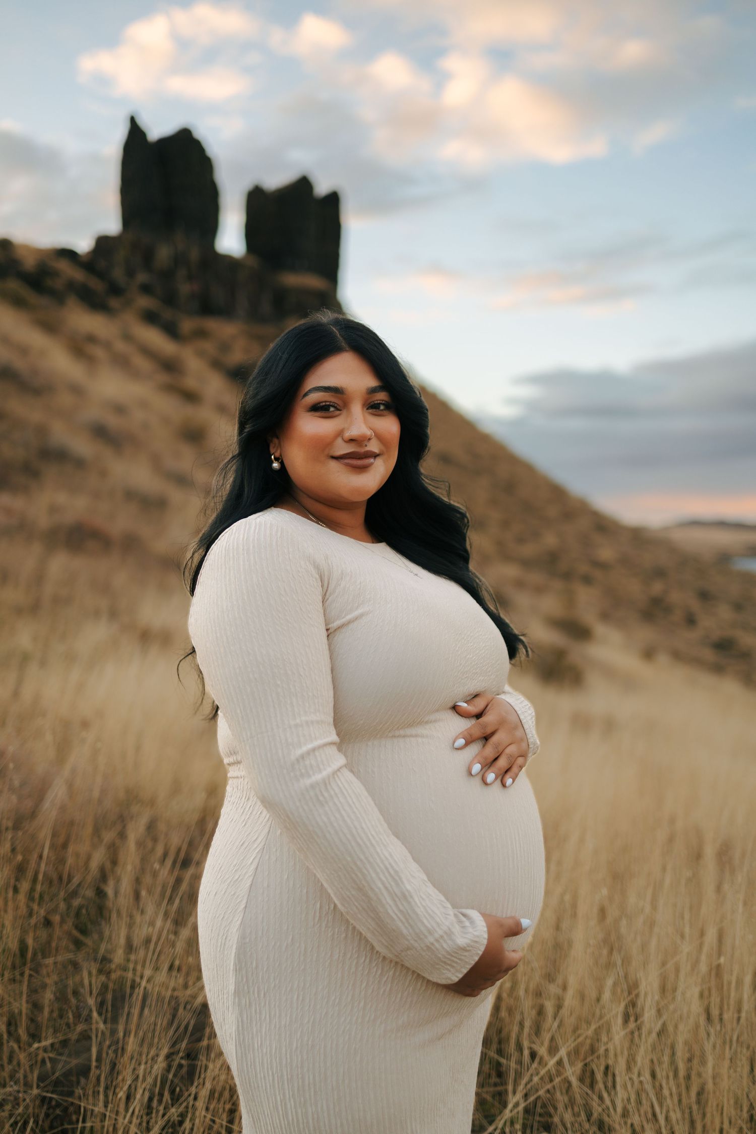 Pregnant woman in cream ribbed dress cradling baby bump at golden hour in scenic landscape.