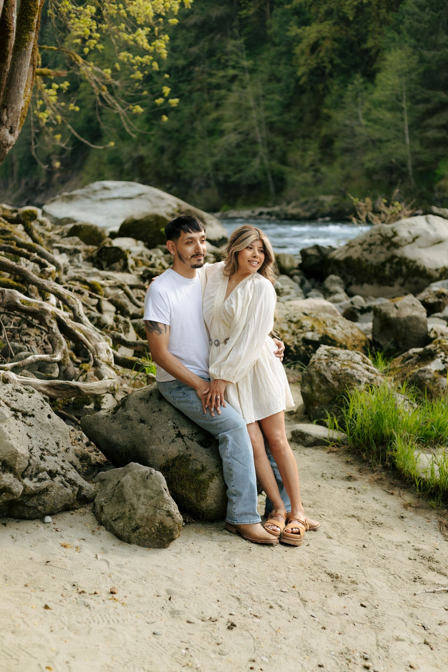 Couple sitting on rocks by a river in a lush forest, man in white tee and jeans, woman in white dress and sandals.