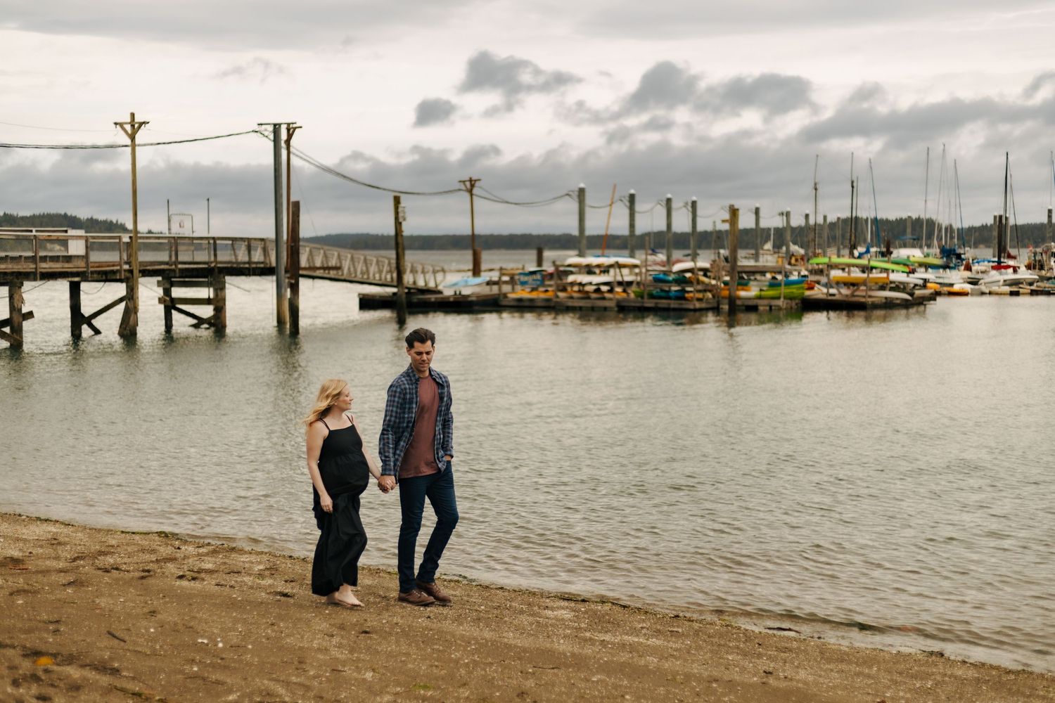Couple holding hands on sandy shore near marina with pier and moored boats on overcast day.
