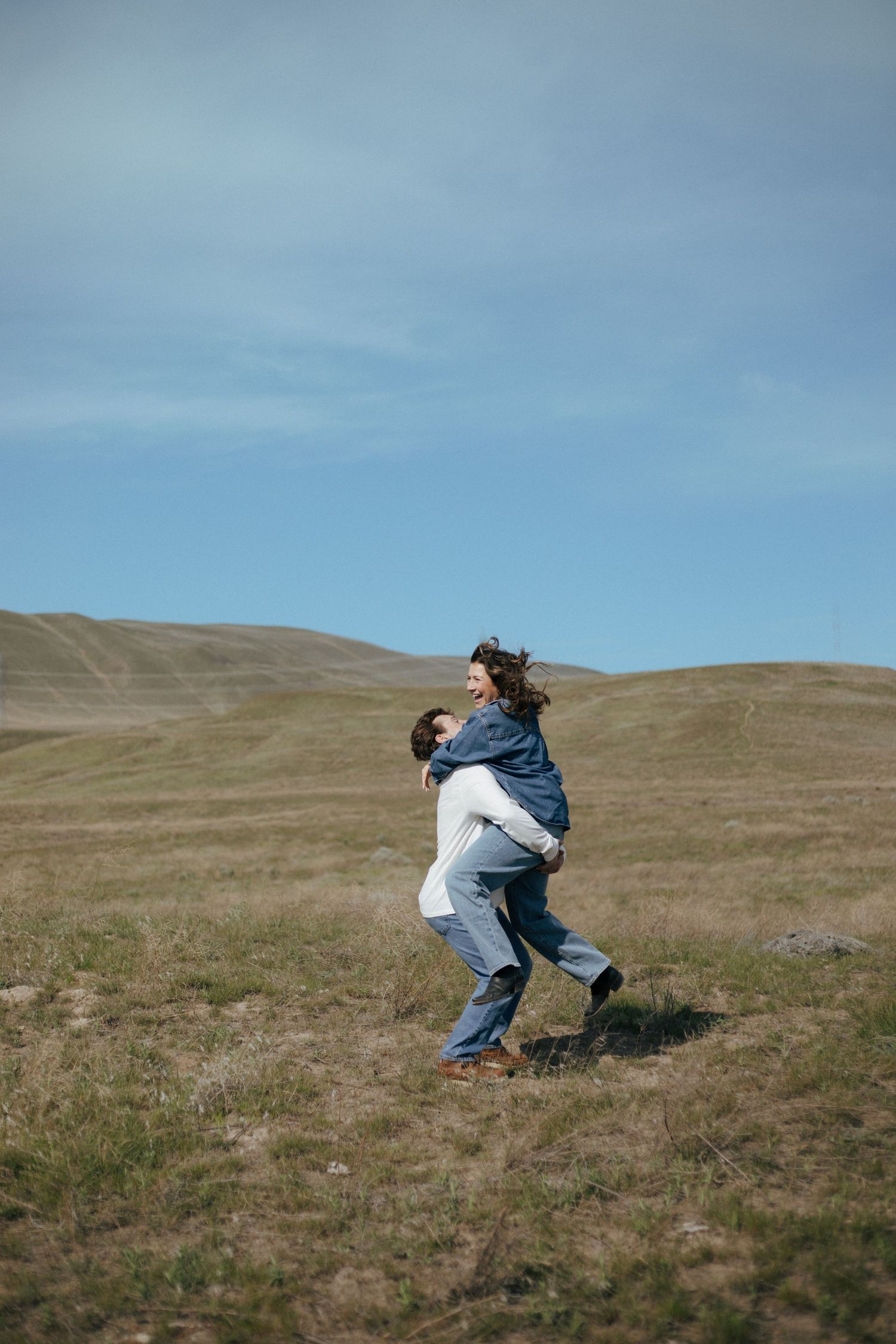 Two figures embrace and spin playfully on a grassy hillside against a bright blue sky on a sunny day.