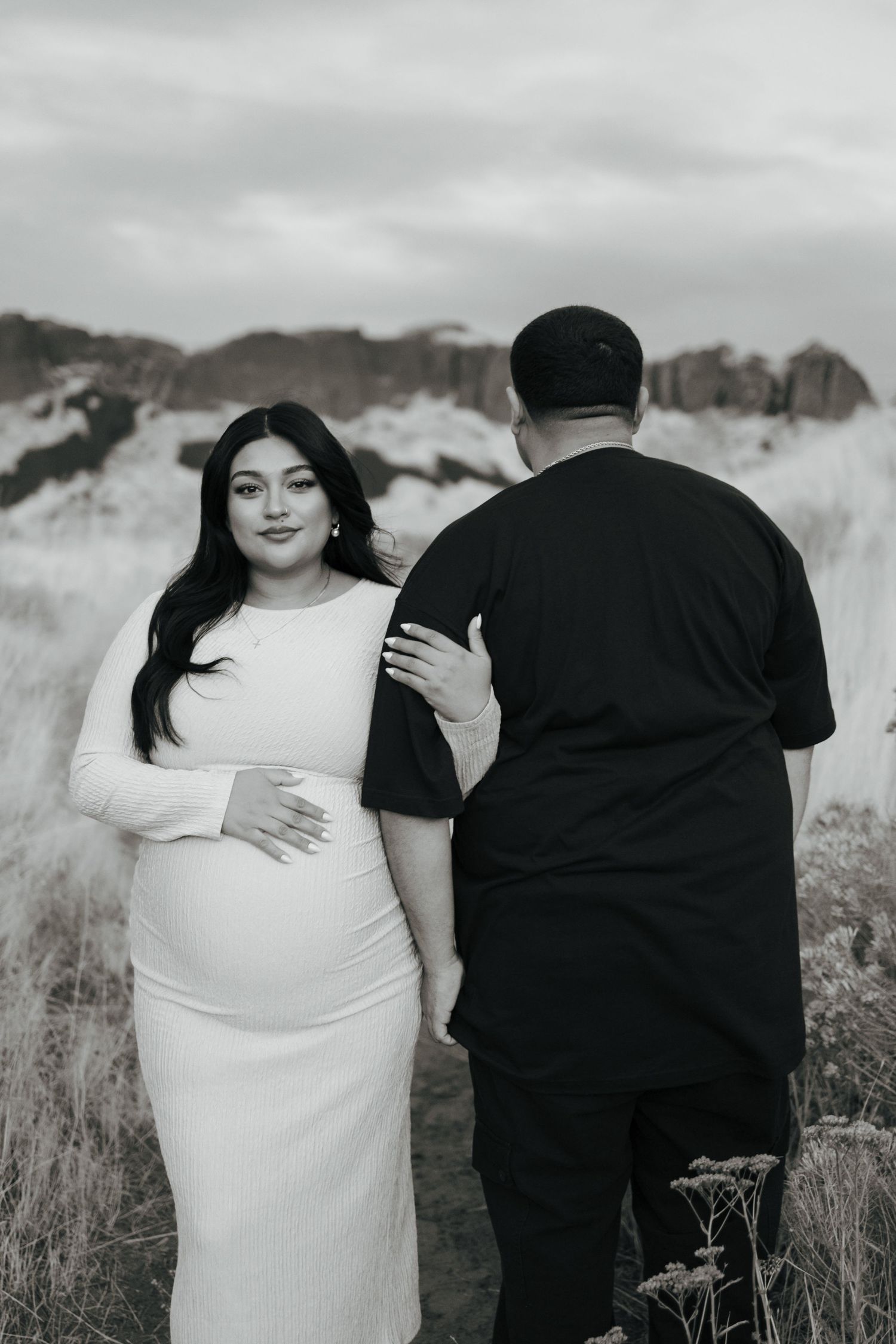 Pregnant woman in white dress poses with partner in black outdoors during maternity photoshoot in natural landscape.