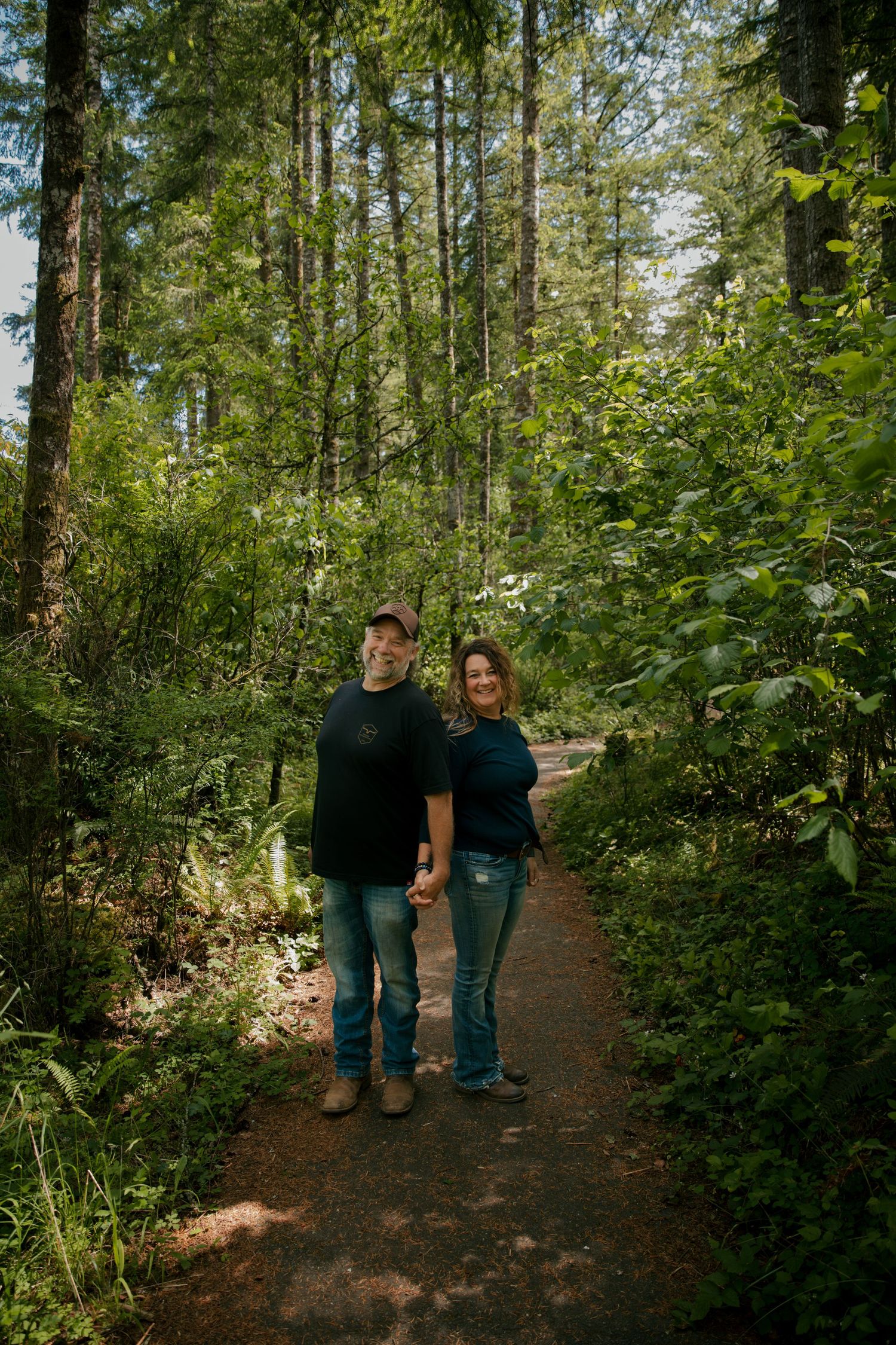A pair stands together on a hiking trail surrounded by lush green forest vegetation and tall evergreen trees.