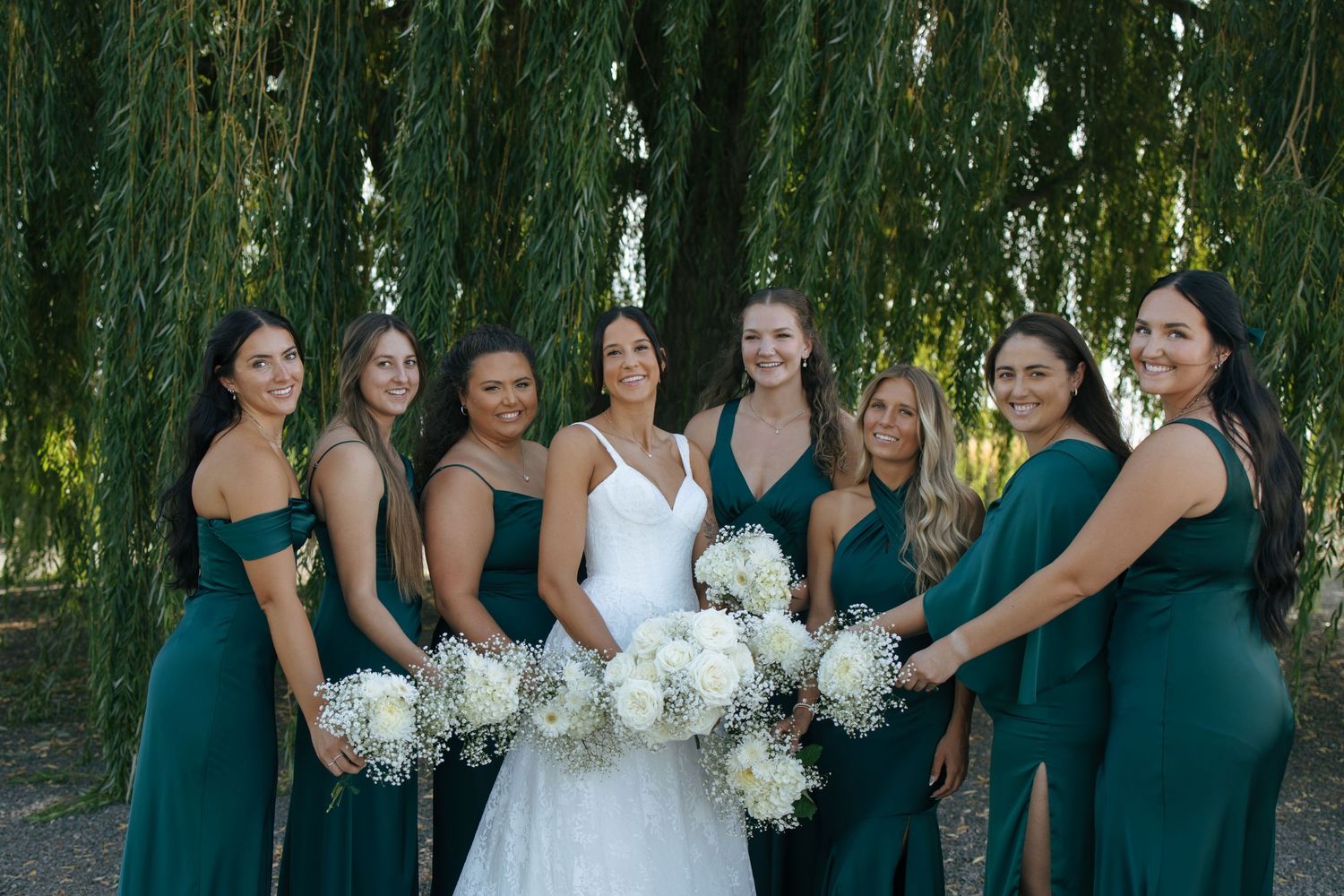 Wedding party in teal dresses poses outdoors under weeping willow trees with their white bouquets.
