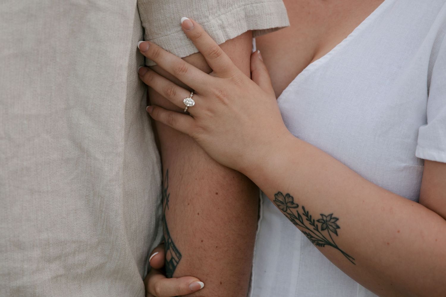 Close-up view of hands with an engagement ring and floral tattoo against white clothing.