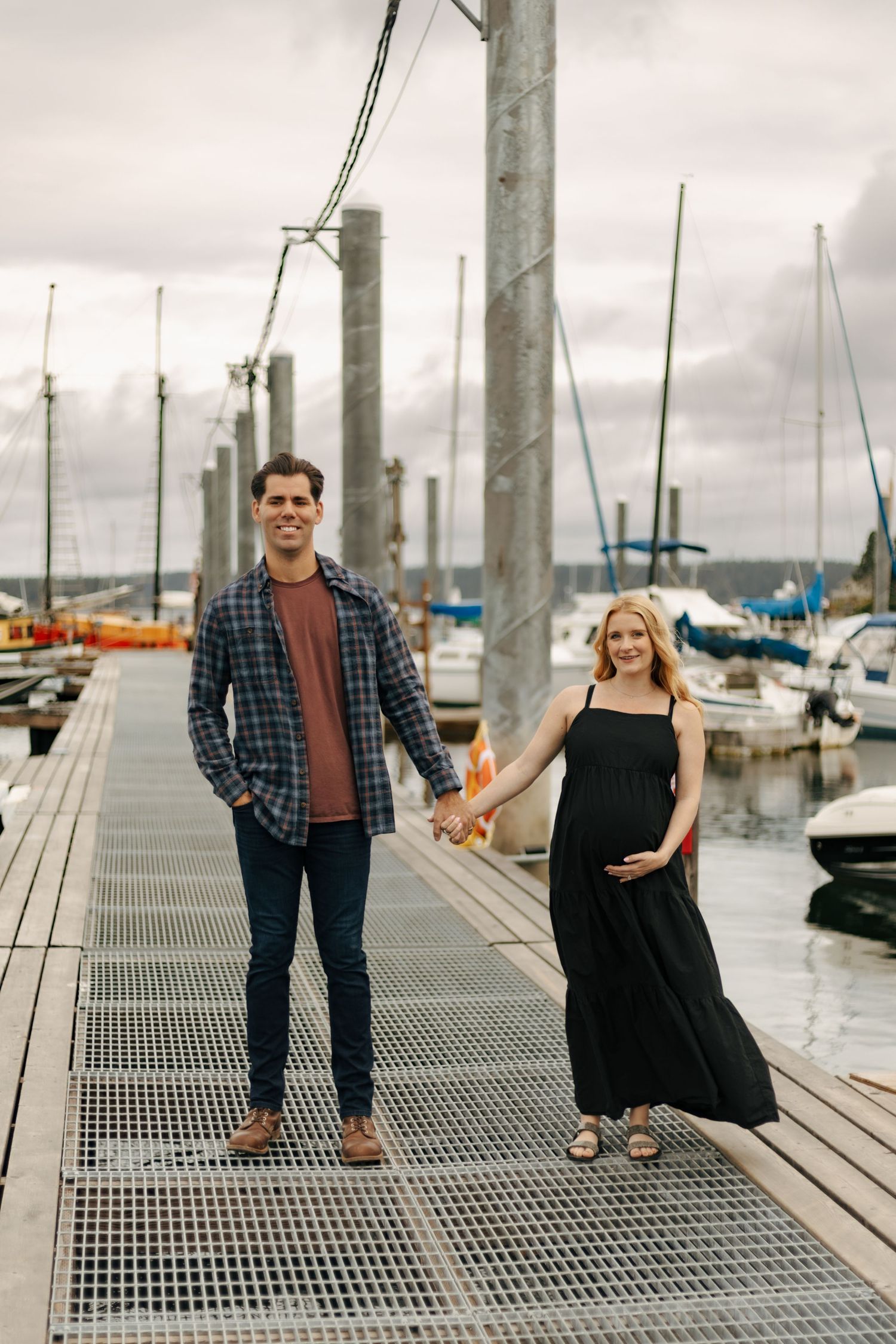 Pregnant couple holding hands on a marina dock with sailboats, man in plaid shirt and woman in black dress.