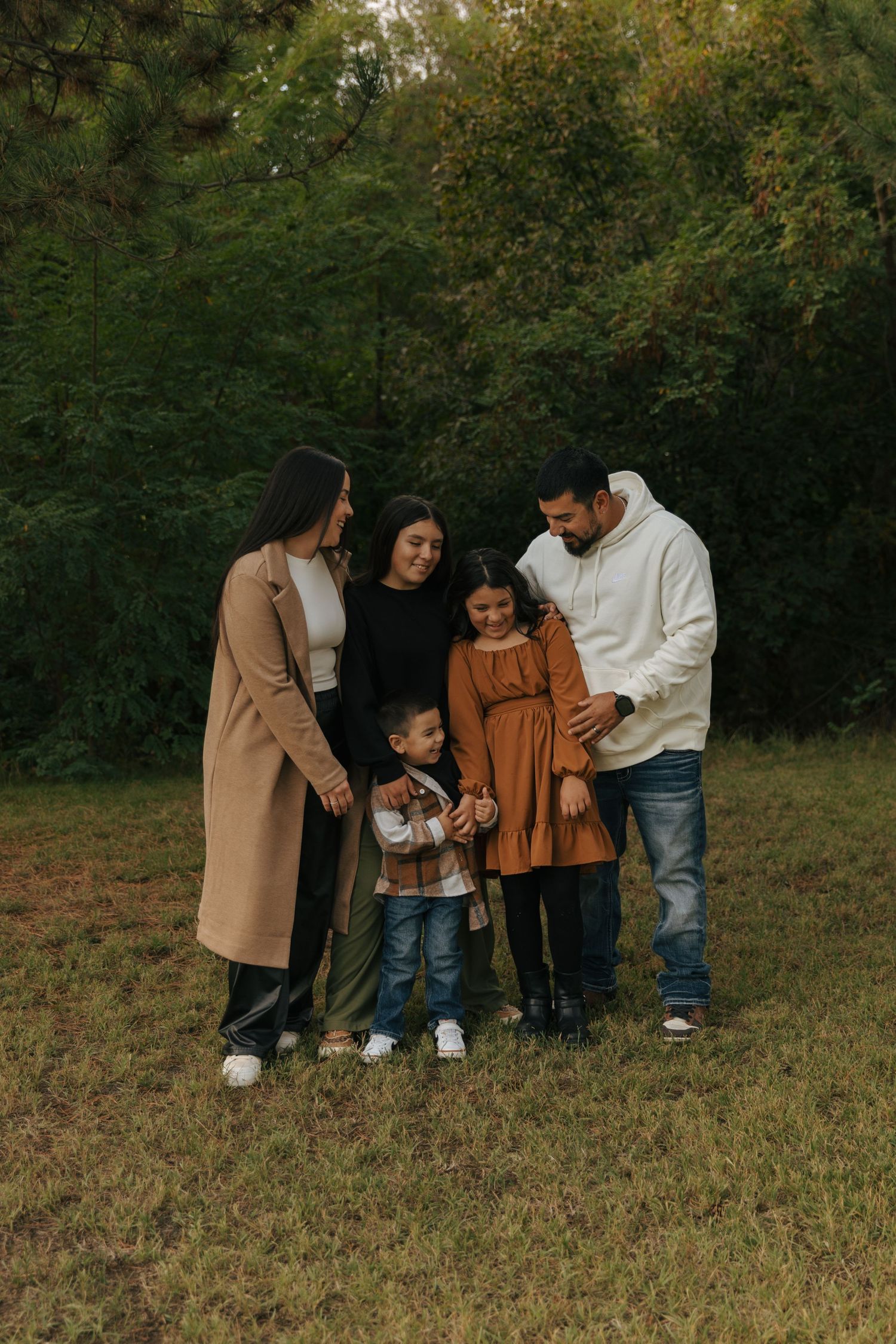 A family wearing coordinated autumn outfits stands together on grass with trees in the background at sunset.