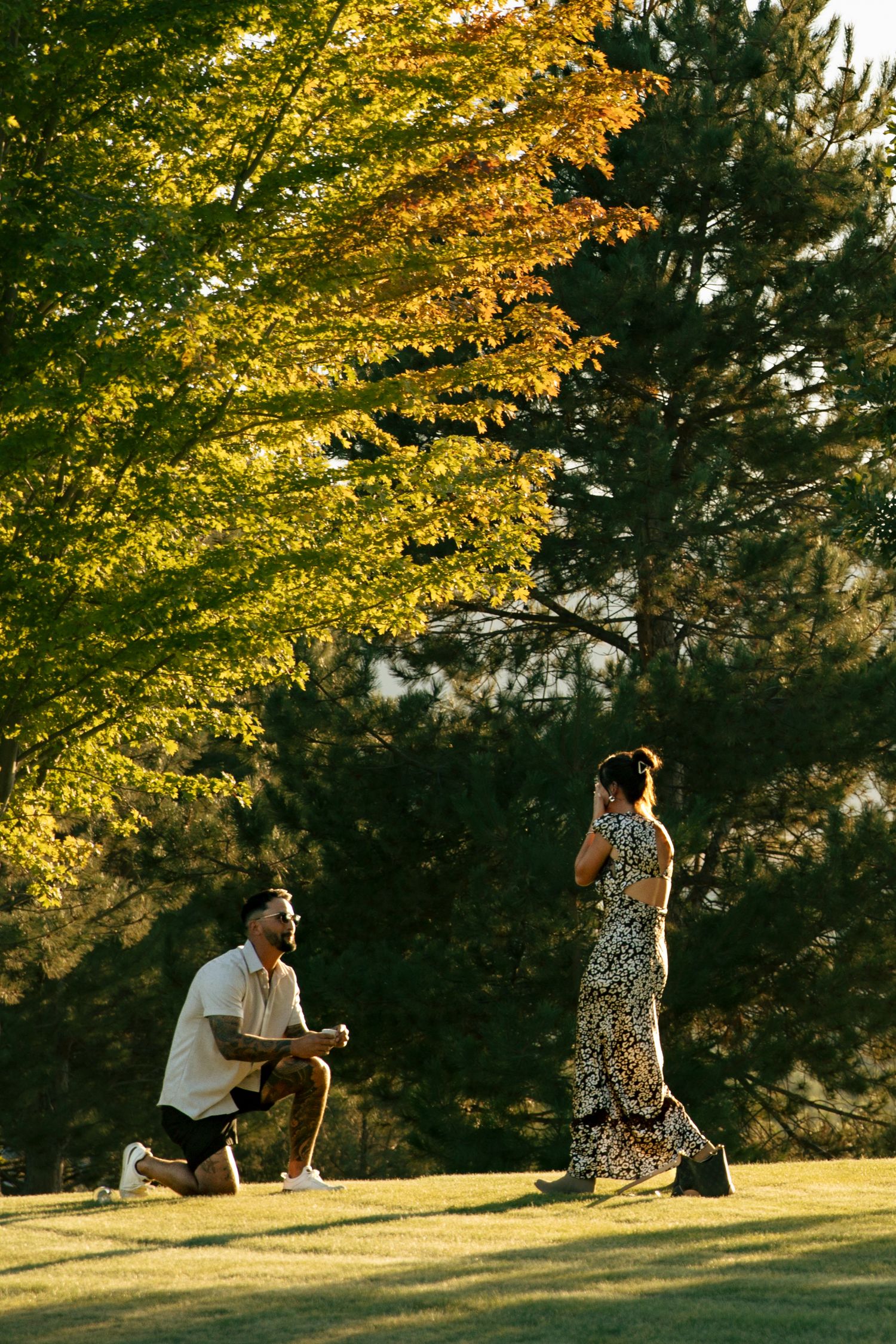 Man kneeling on one knee proposing to woman in floral dress outdoors among autumn trees at golden hour.