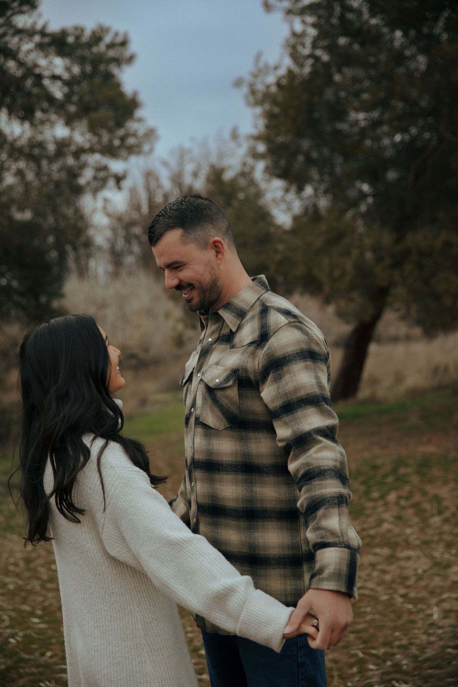 Couple holding hands outdoors in autumn setting with person in plaid flannel and other in cream sweater.