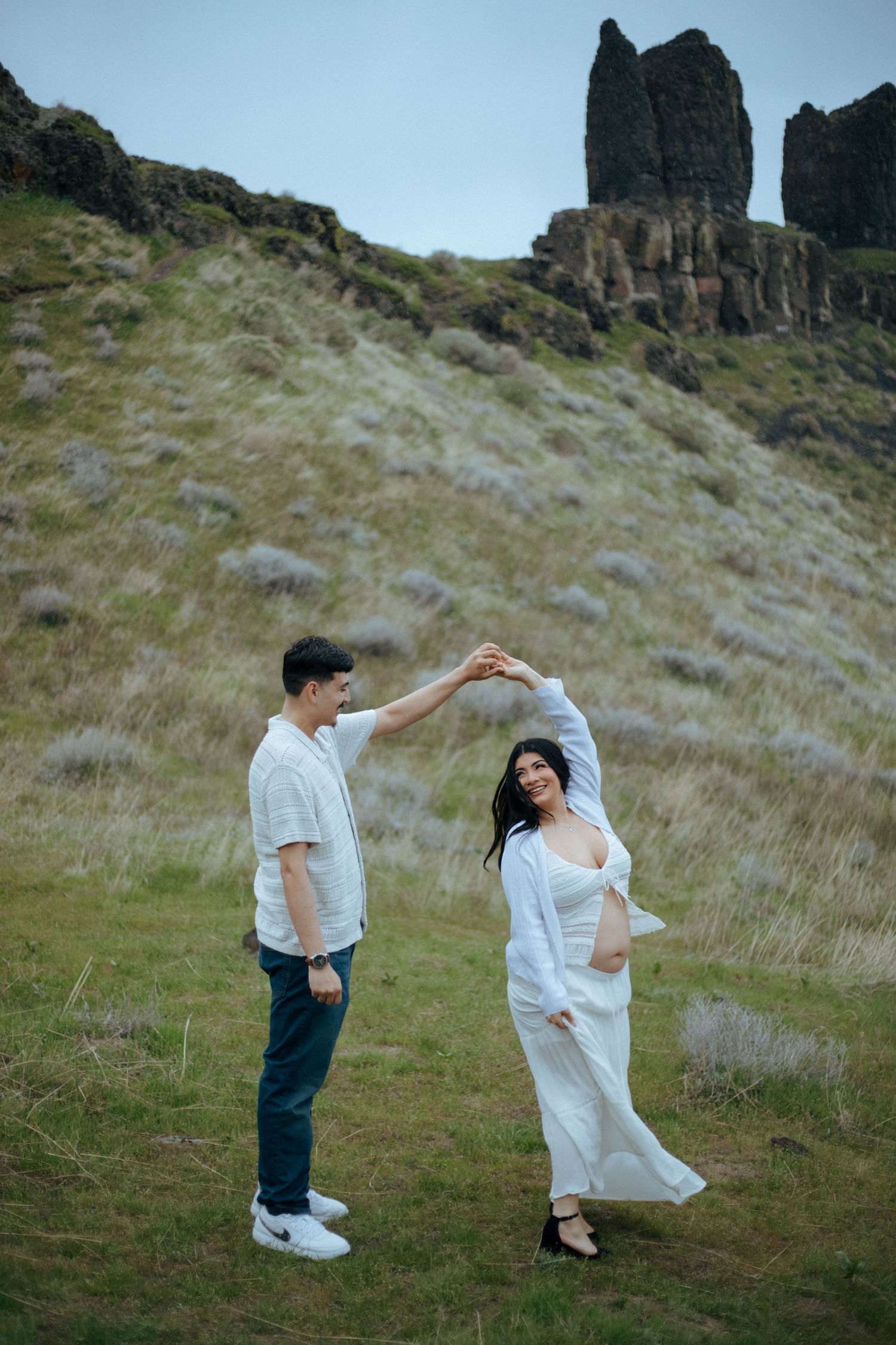 A couple dances together against a backdrop of scenic rocky cliffs in a grassy meadow with blue sky overhead.