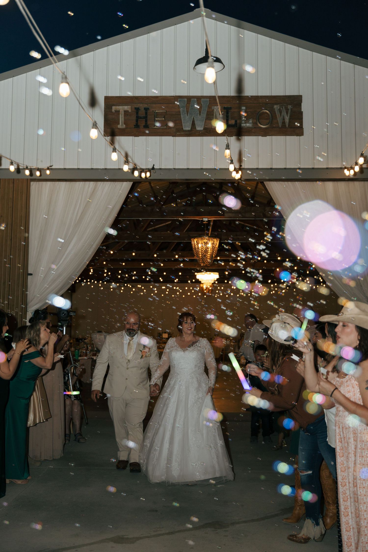 Bride and groom exit The Willow venue surrounded by bubbles and glowing wands held by cheering guests at night.