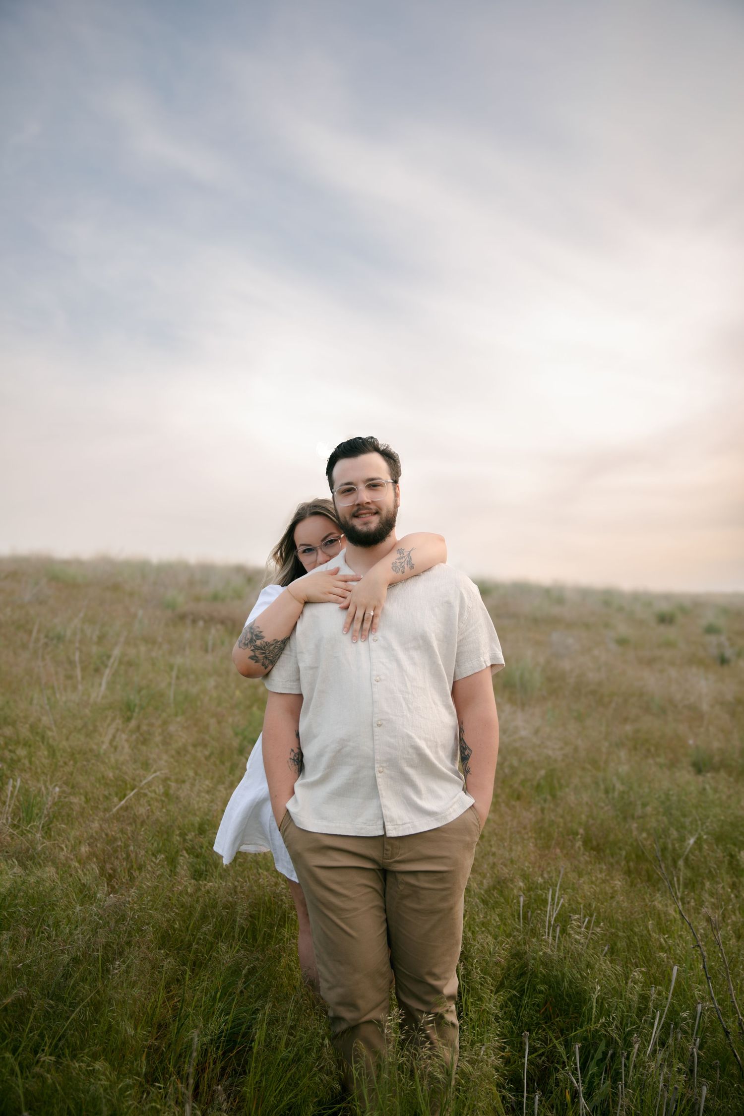 A couple embraces while standing in a grassy meadow during sunset with moody cloudy skies overhead.