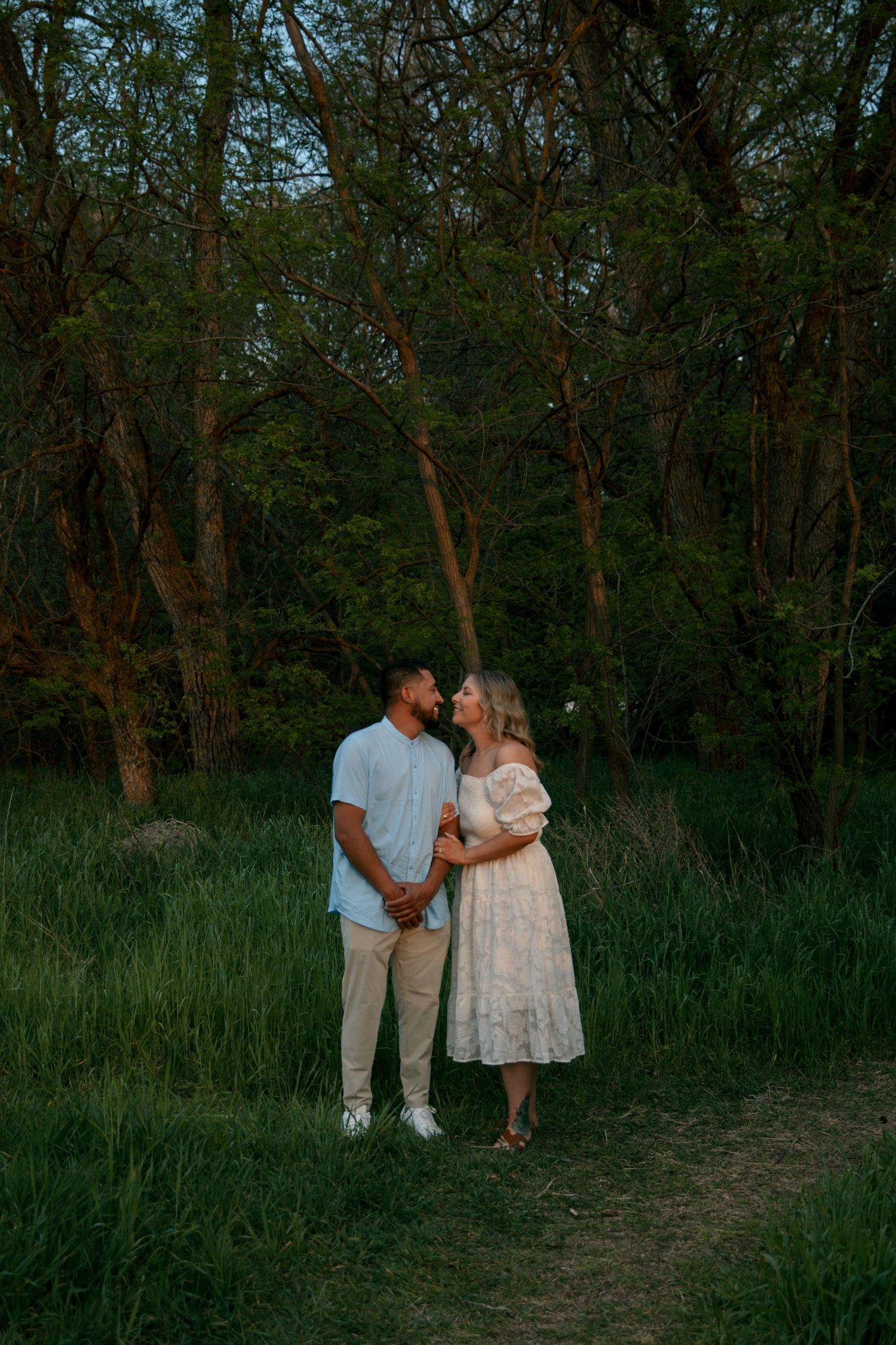 A couple stands together on a path through tall grass and woods during golden hour at sunset.