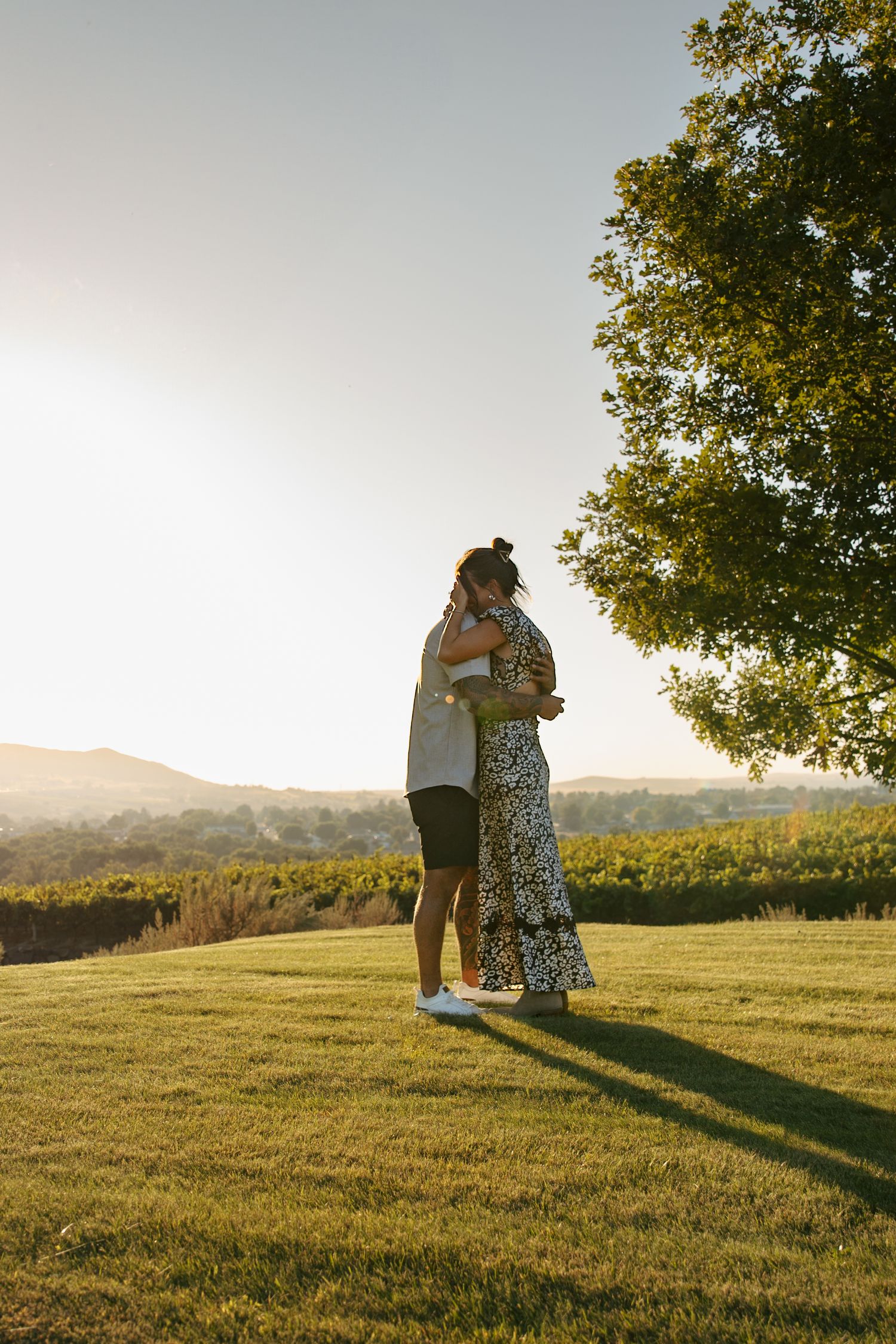 Couple embracing at golden hour on green lawn with scenic vineyard and tree backdrop.