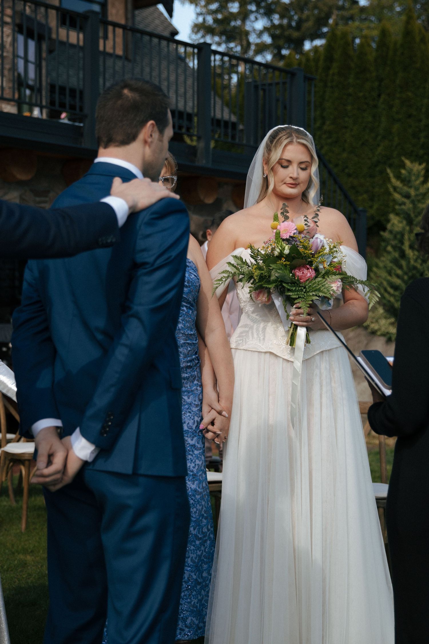 Bride in white gown holding floral bouquet stands with groom in blue suit during outdoor wedding ceremony.
