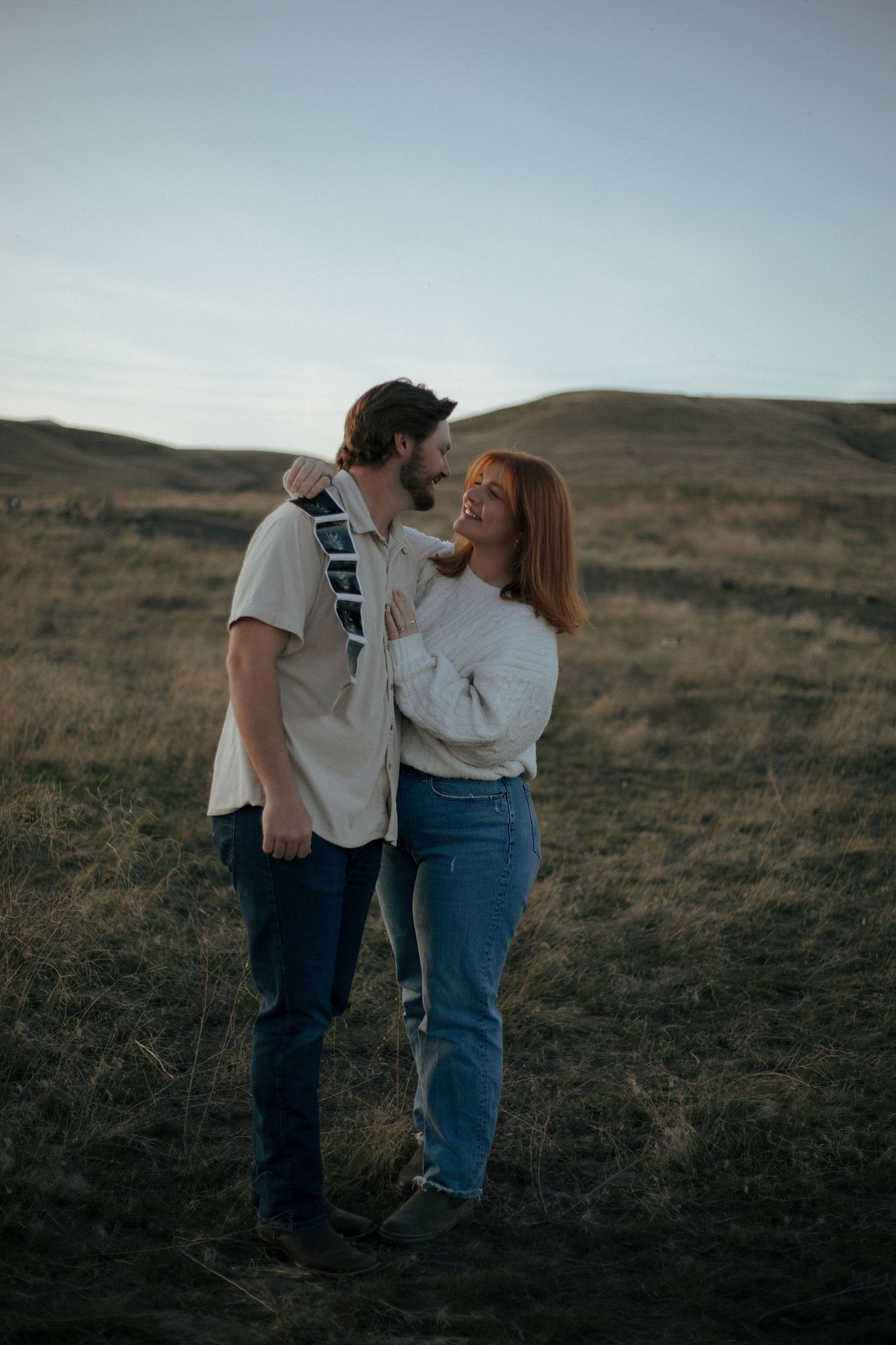 A couple shares an intimate moment in a field at sunset against rolling hills in the background.