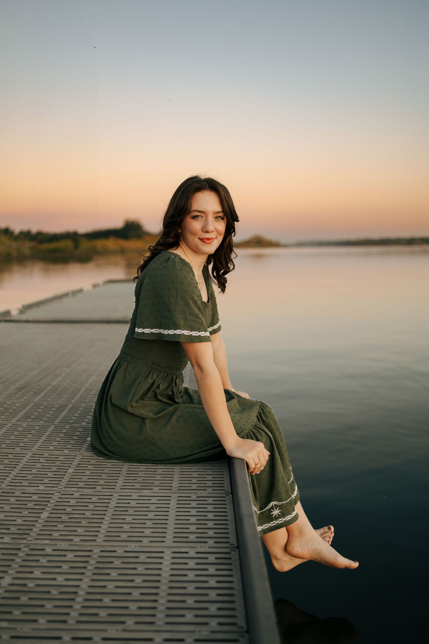 Person in dark clothing sits on a concrete ledge near a tranquil lake at sunset creating a peaceful silhouette.