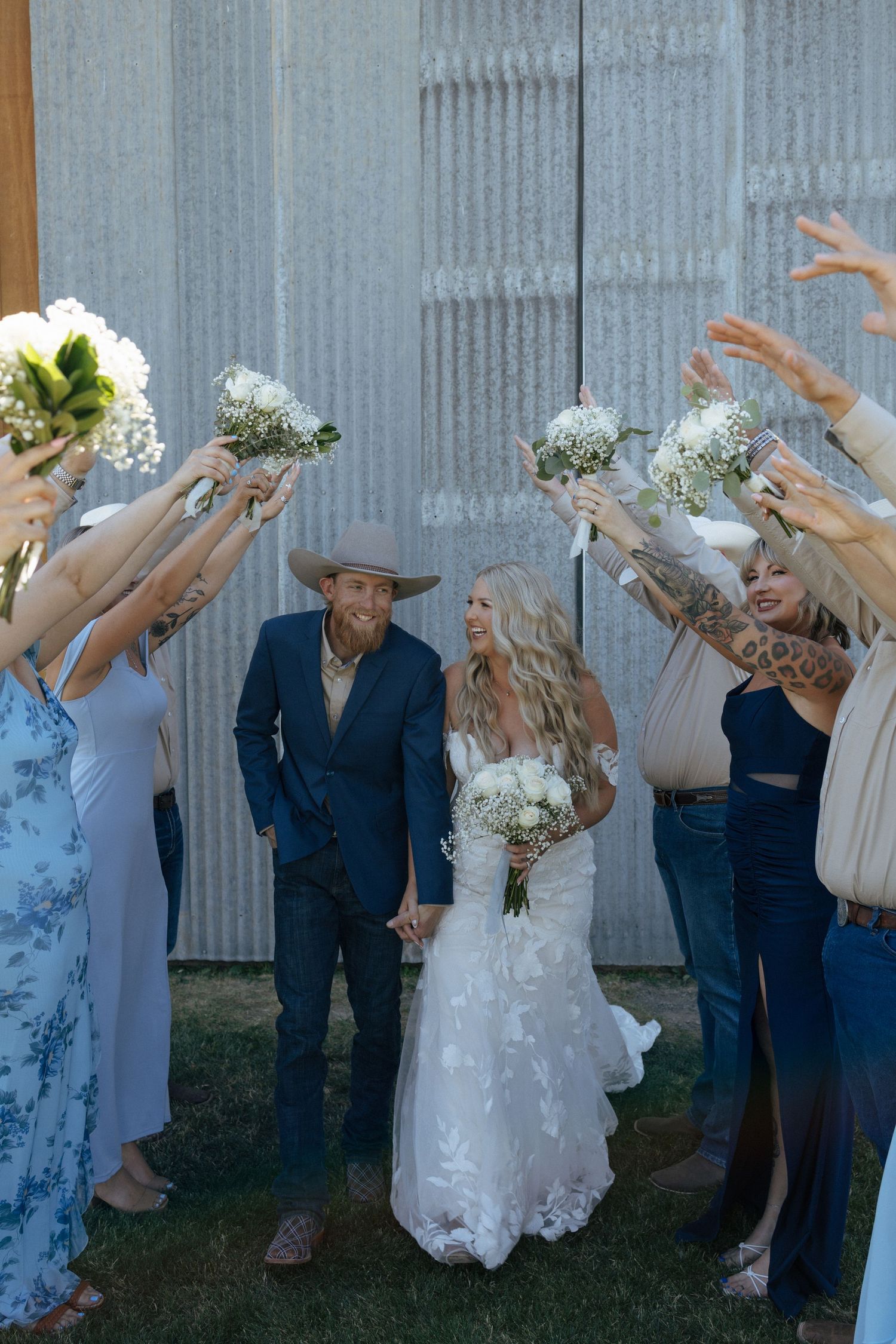 Bride and groom walk through bouquet arch with wedding party at rustic barn venue.