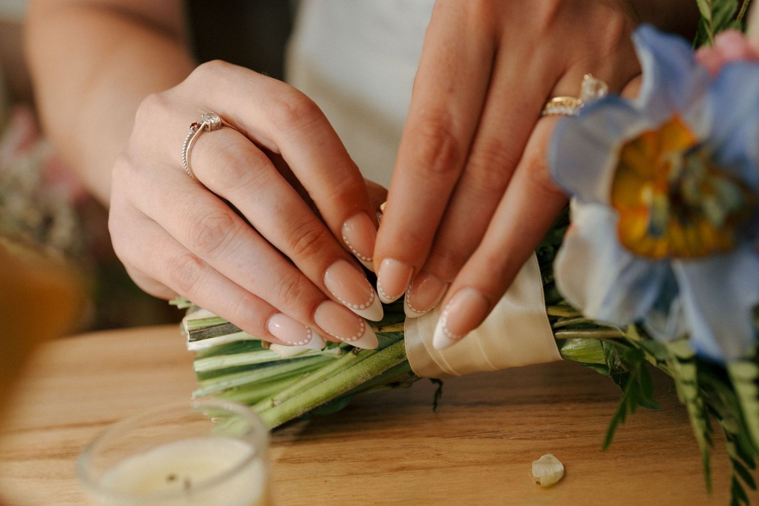 Close-up of hands with rings arranging a floral bouquet on a wooden surface with blue flowers.