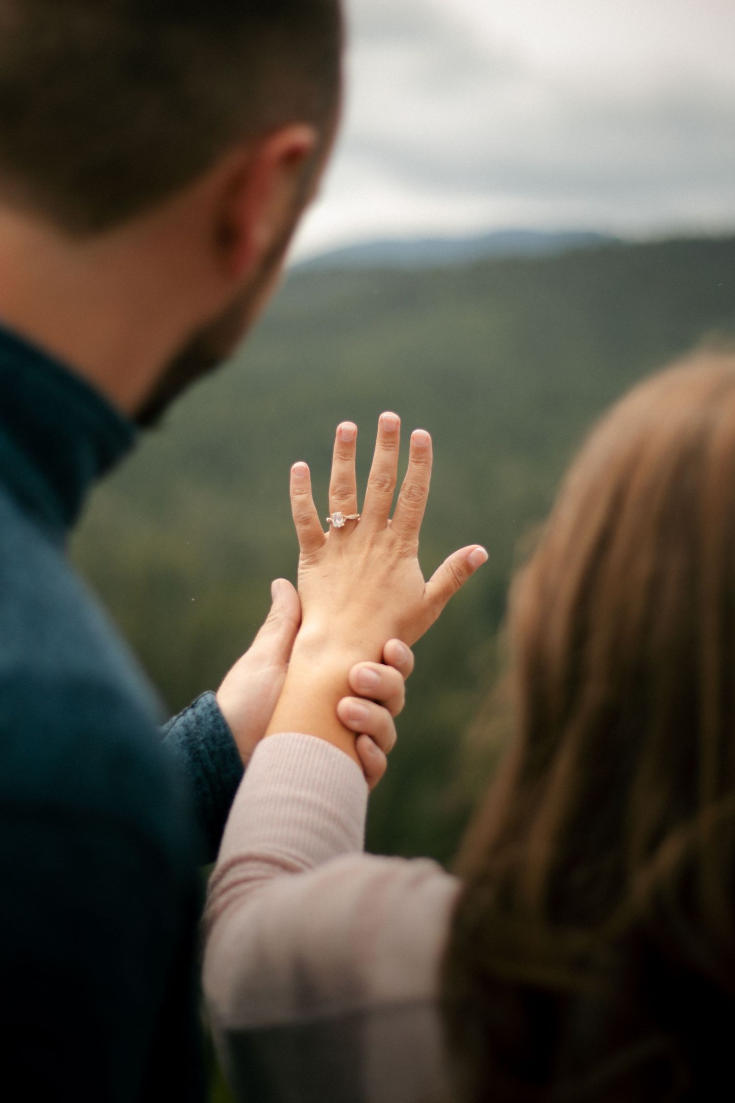 Two people holding hands and showing off an engagement ring against a scenic mountain landscape backdrop.