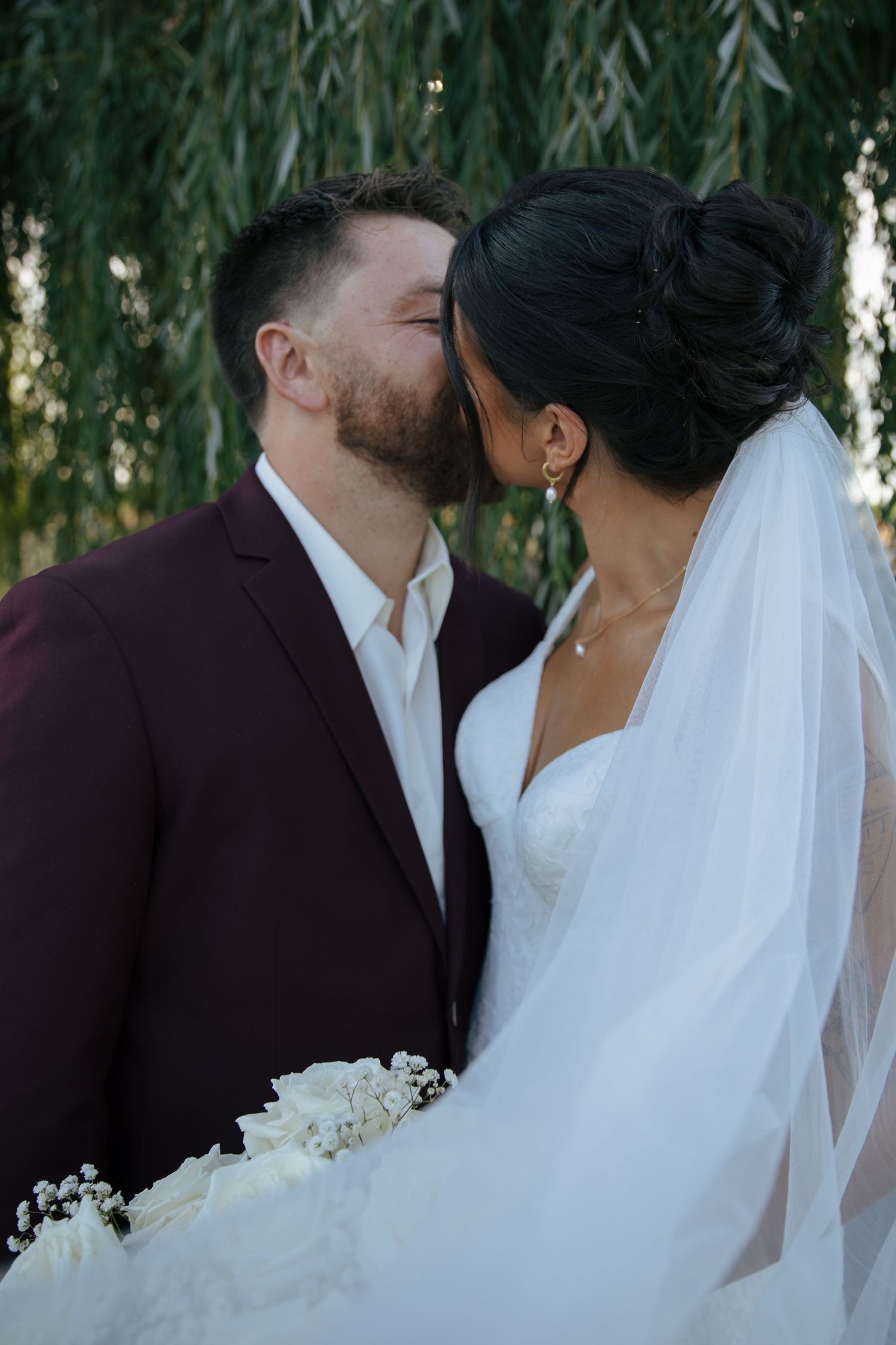 A romantic wedding portrait under a weeping willow tree with flowing veil and white bouquet.