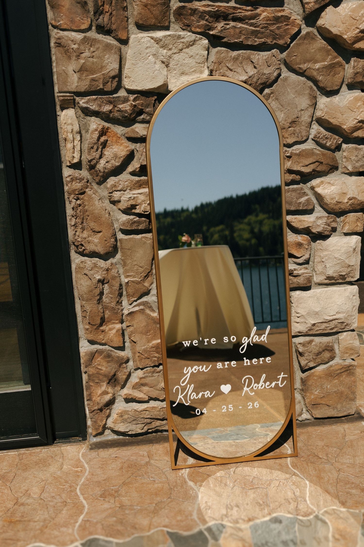 Gold-framed arch mirror with wedding welcome message for Klara and Robert dated 04-25-26 against stone wall.