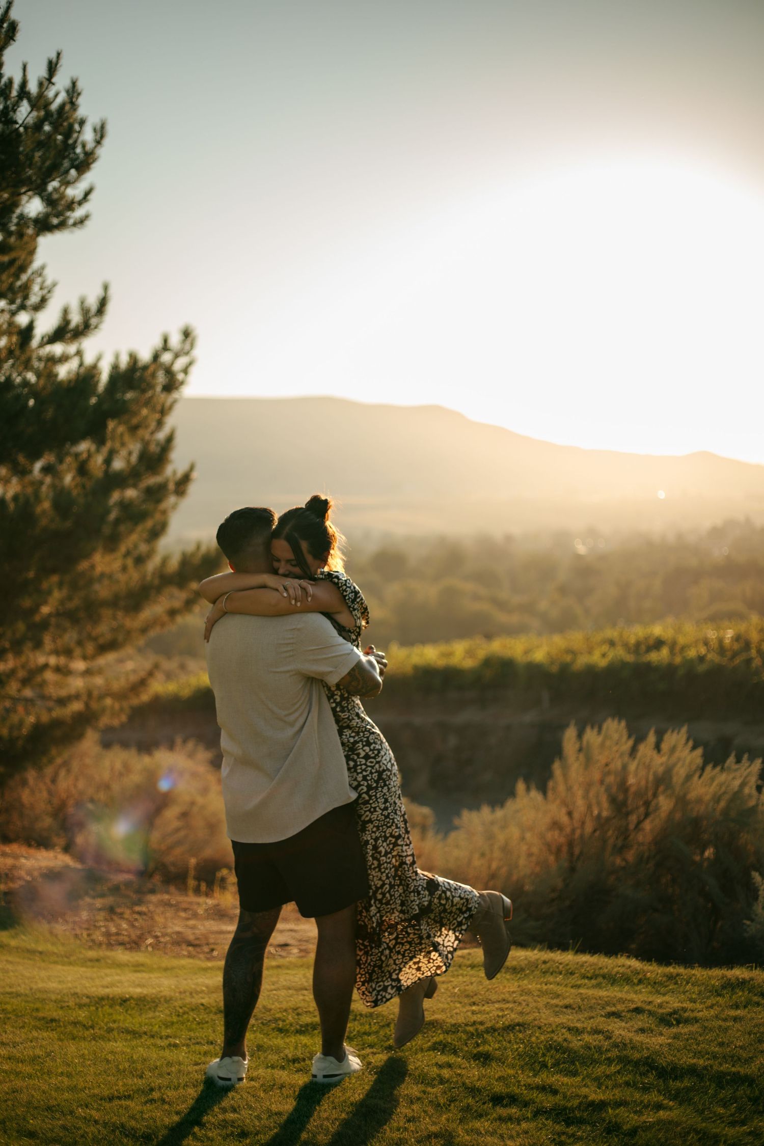 Couple embracing at golden hour sunset with mountain landscape and lush greenery in the background.
