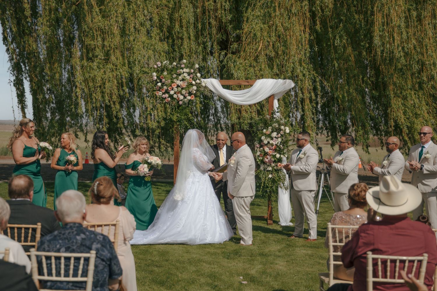 Outdoor wedding ceremony under a white draped arch with bride, groom, and green-gowned bridesmaids beside a weeping willow tree.