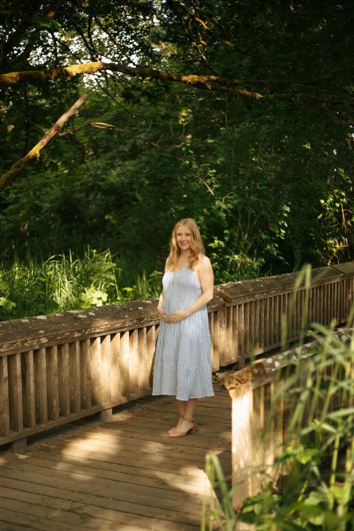 A person in a light blue summer dress stands on a wooden bridge walkway surrounded by lush green foliage.