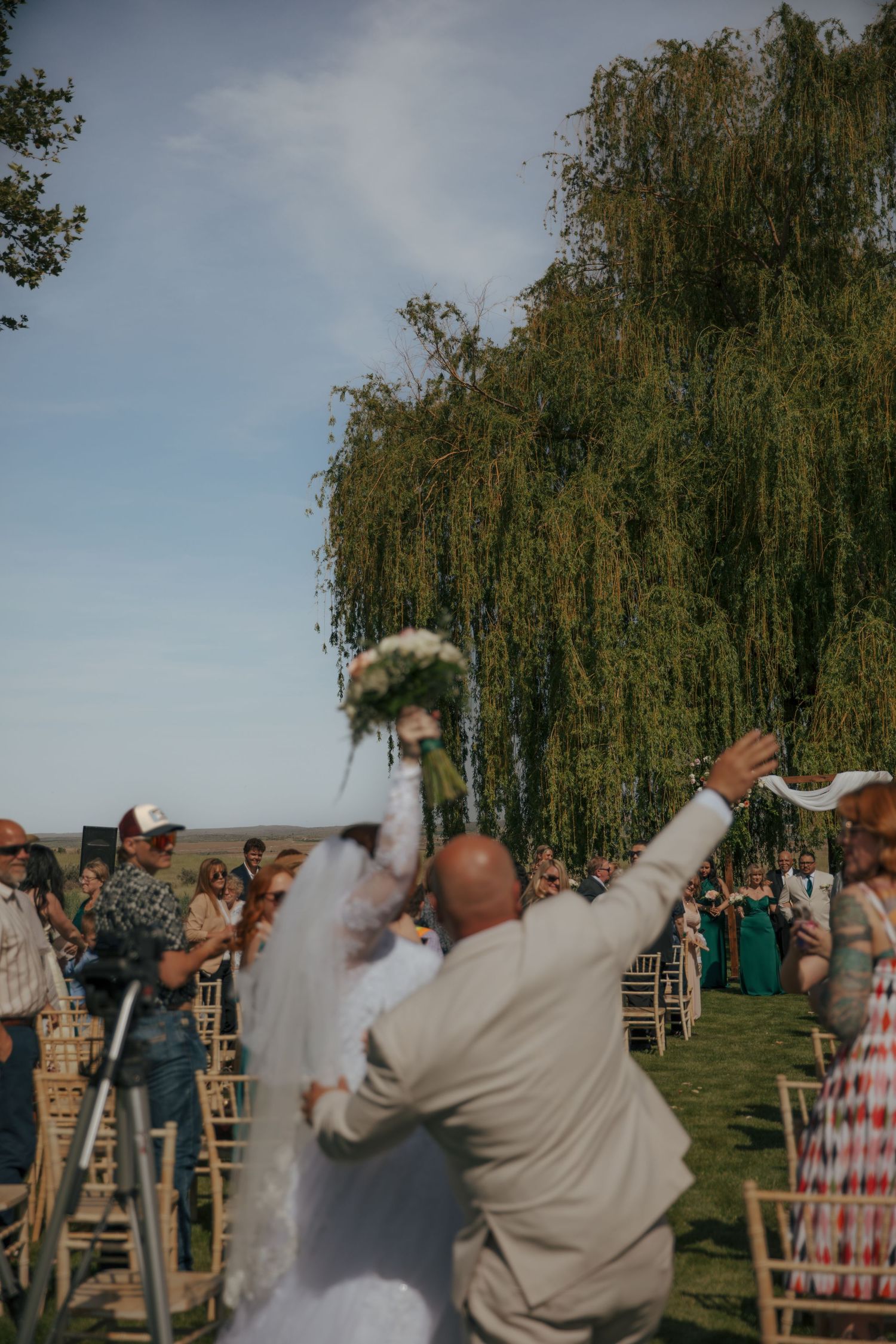 Bride raises bouquet joyfully as groom lifts her at outdoor wedding ceremony near weeping willow tree.