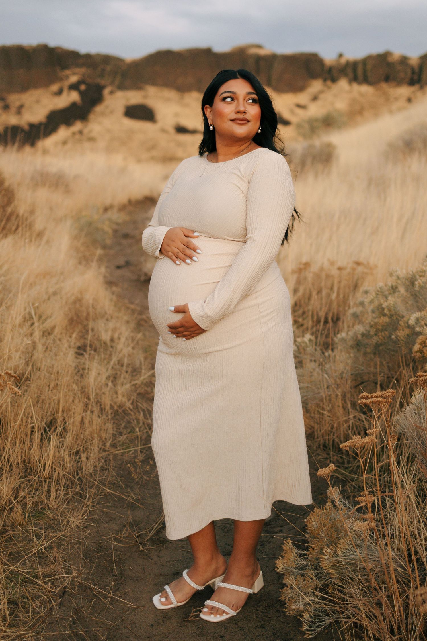 Expectant mother in white dress poses in golden field during sunset maternity photoshoot.
