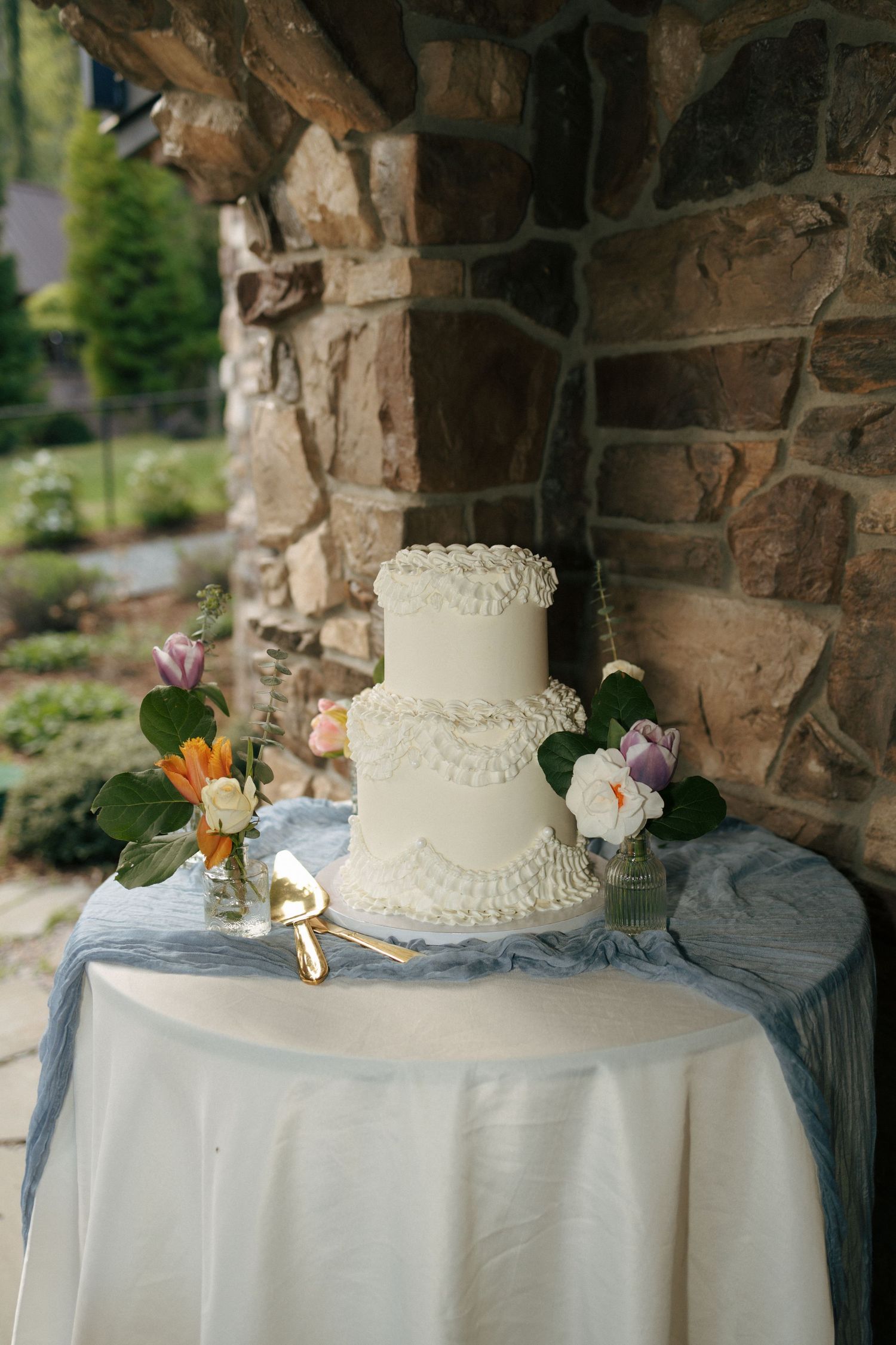 Elegant two-tier white wedding cake with ruffled frosting on a draped table, flanked by floral arrangements outdoors.