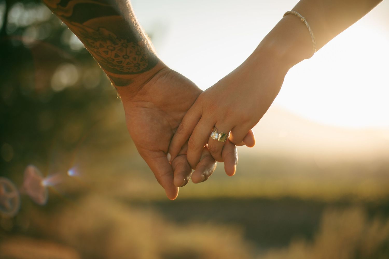 Couple holding hands outdoors at sunset, one with tattoos and one wearing a diamond ring, with a blurred nature background.