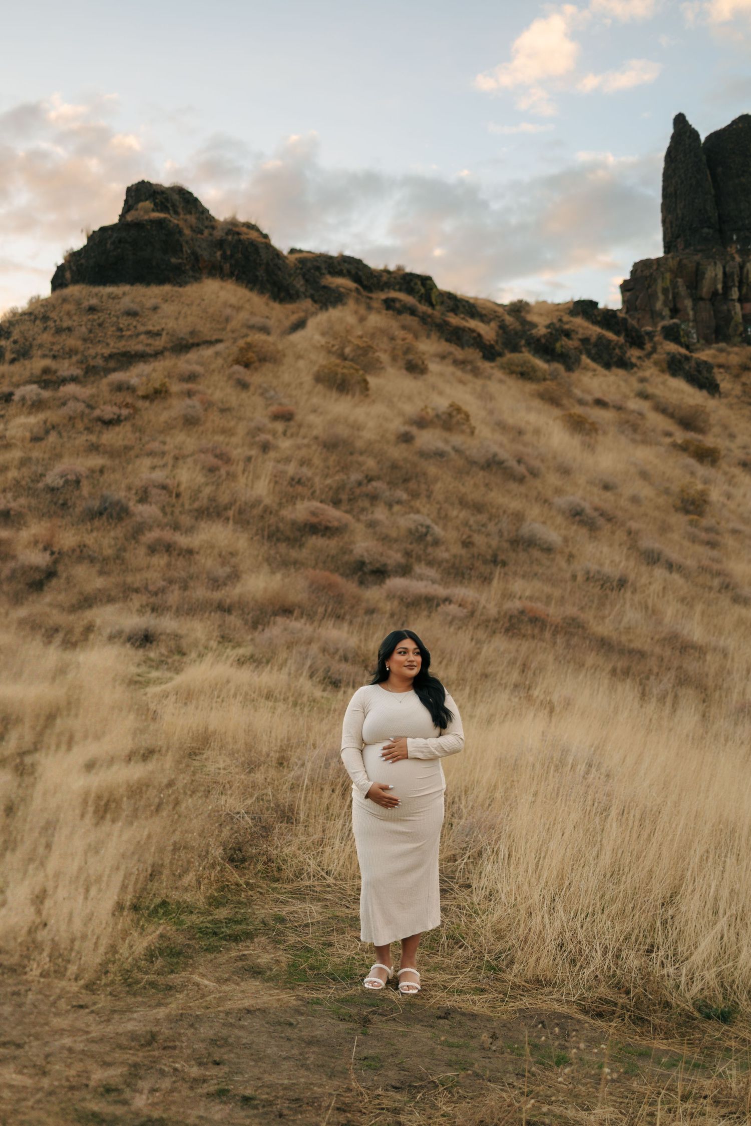 Pregnant woman in white dress cradling belly in golden field with rocky hills at sunset.