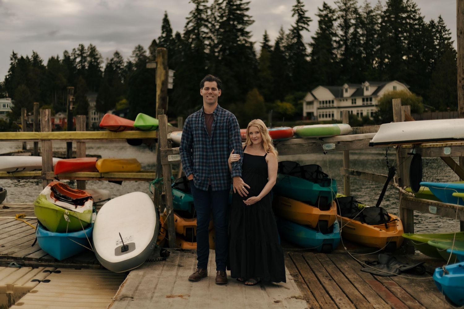 Couple posing on a wooden dock surrounded by colorful kayaks and lush trees in a scenic waterfront setting.
