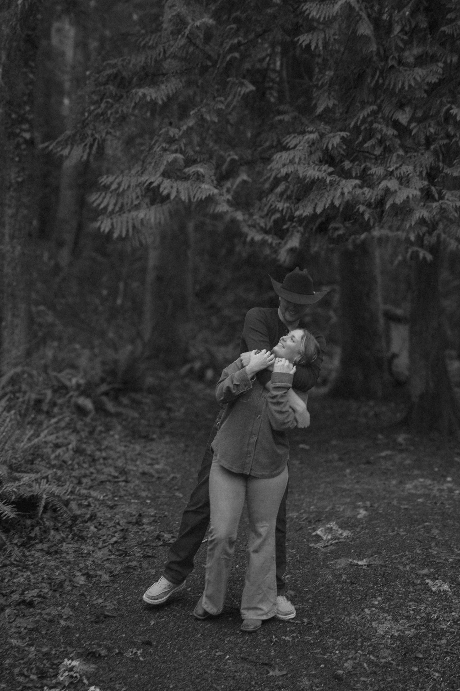 A couple shares an intimate embrace in a wooded forest setting captured in black and white photography.