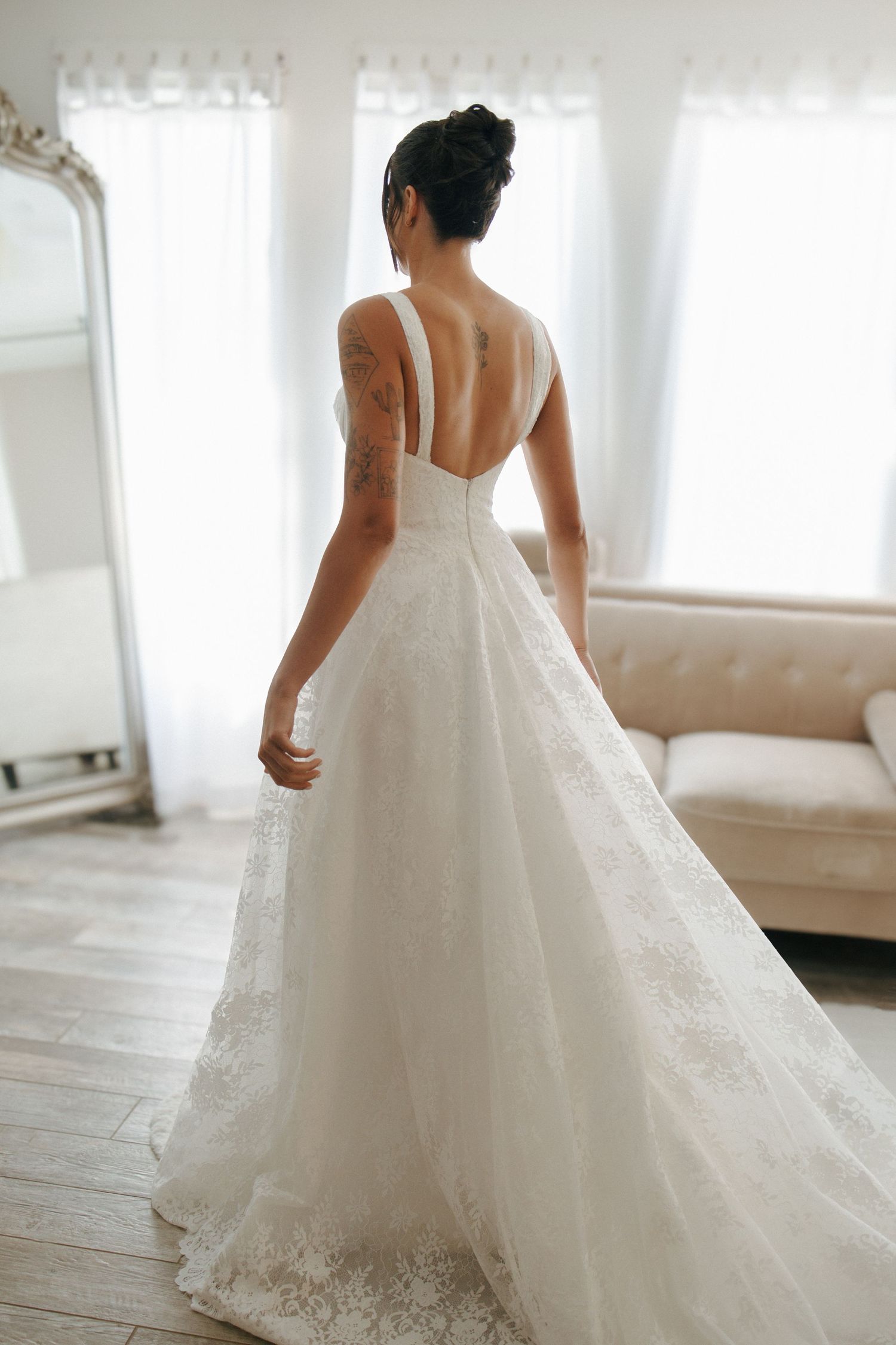 A flowing white wedding dress with intricate lace details is shown from multiple angles on a wood floor in soft light.