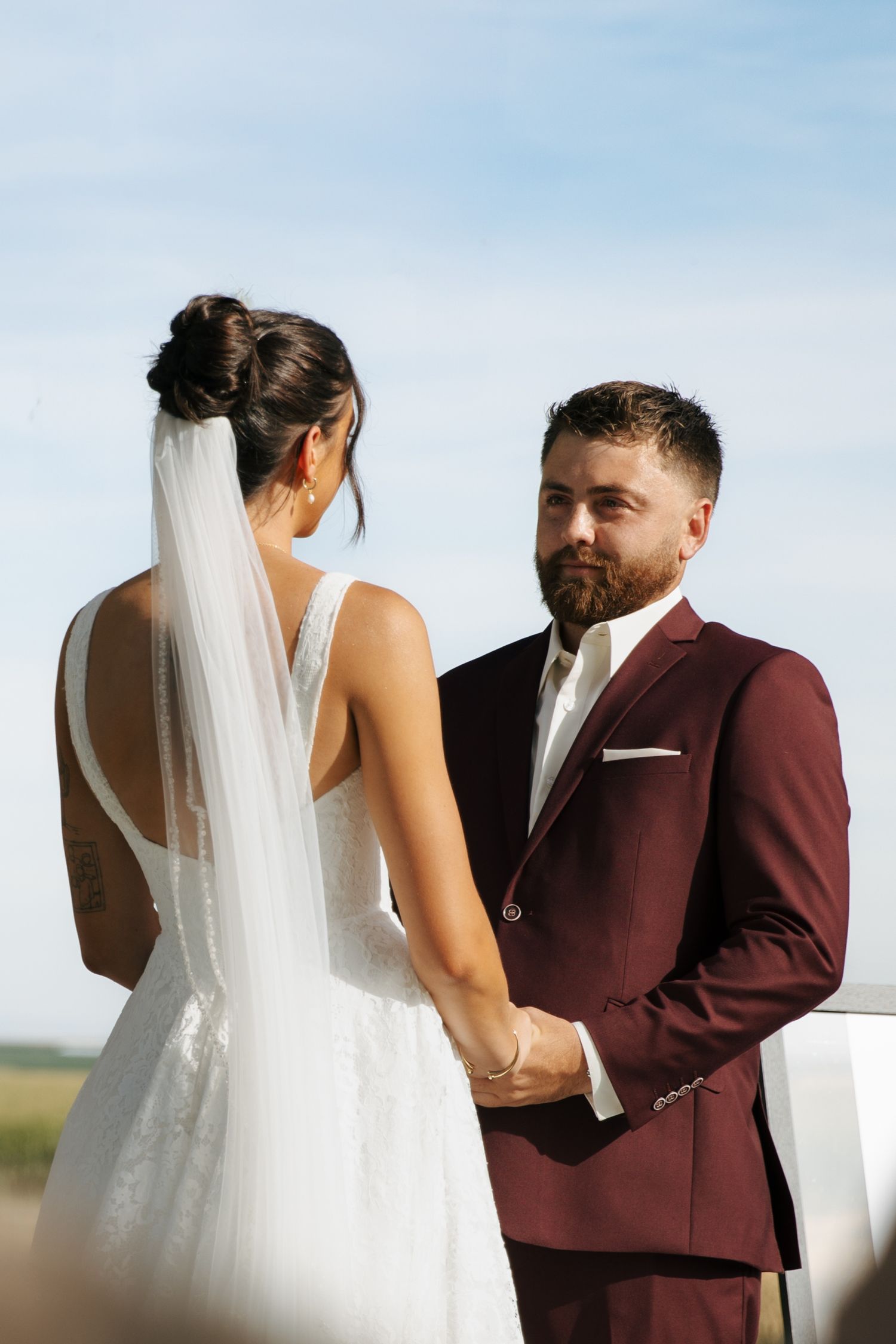A couple exchanges vows during an outdoor wedding ceremony, with the bride in white and groom in burgundy suit.