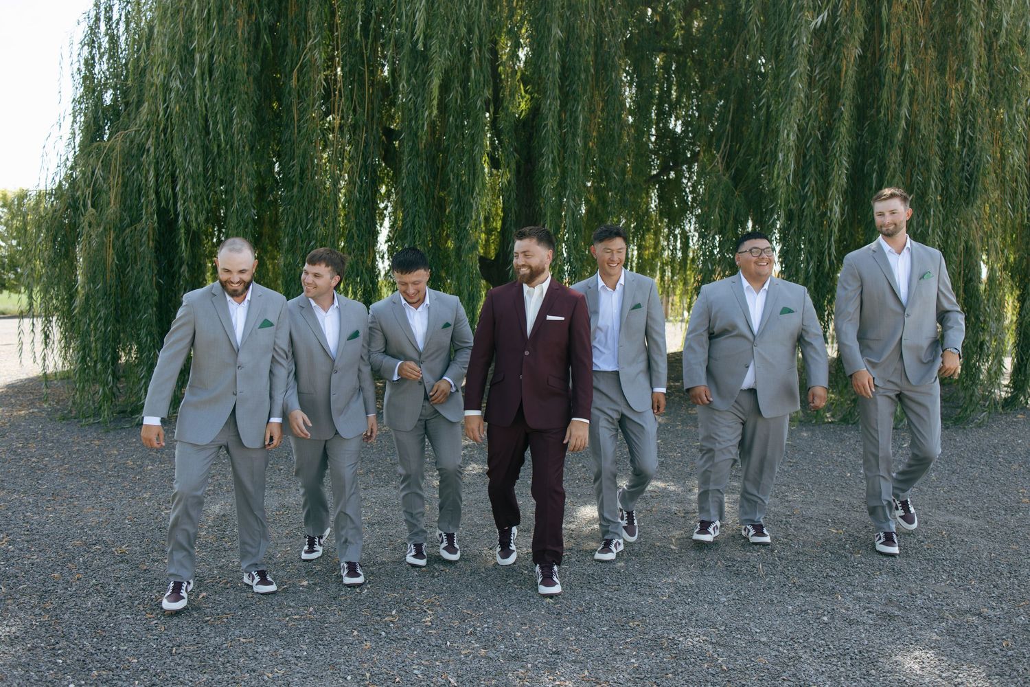 A sequence of people in formal attire walking along a path lined with drooping willow trees on a sunny day.