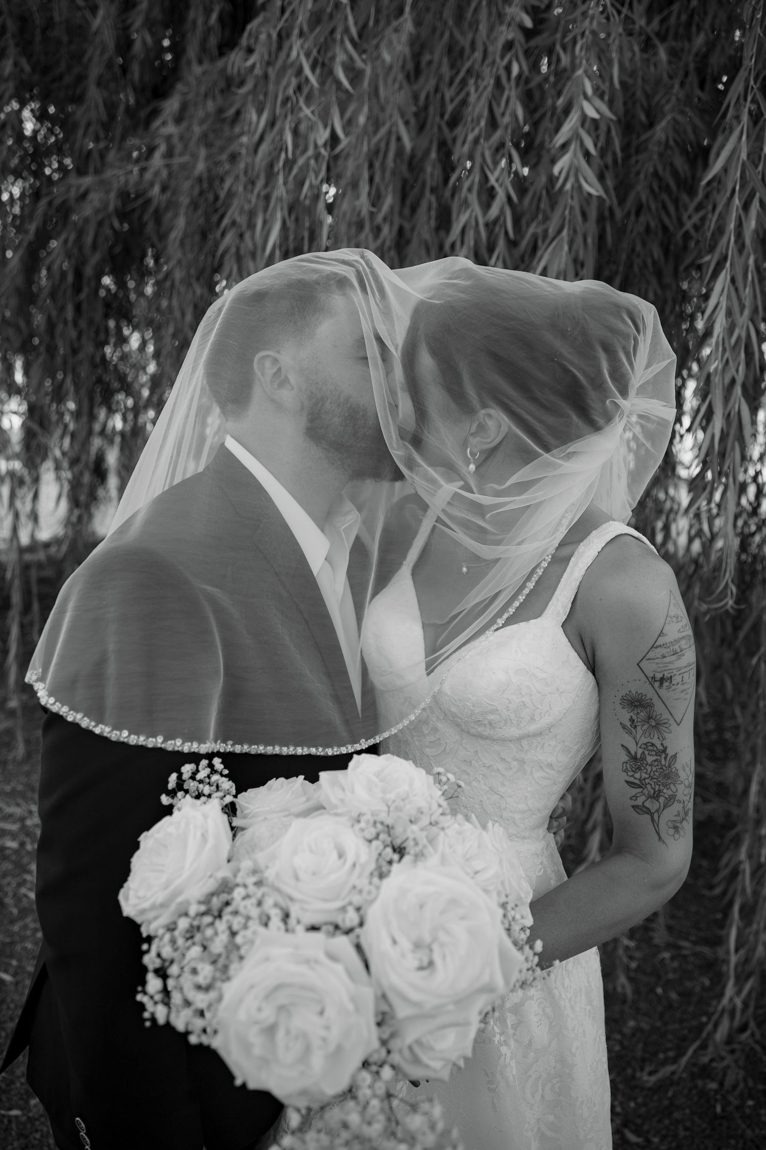 Romantic black and white wedding photo of a couple kissing under a veil beneath a willow tree with white rose bouquet.