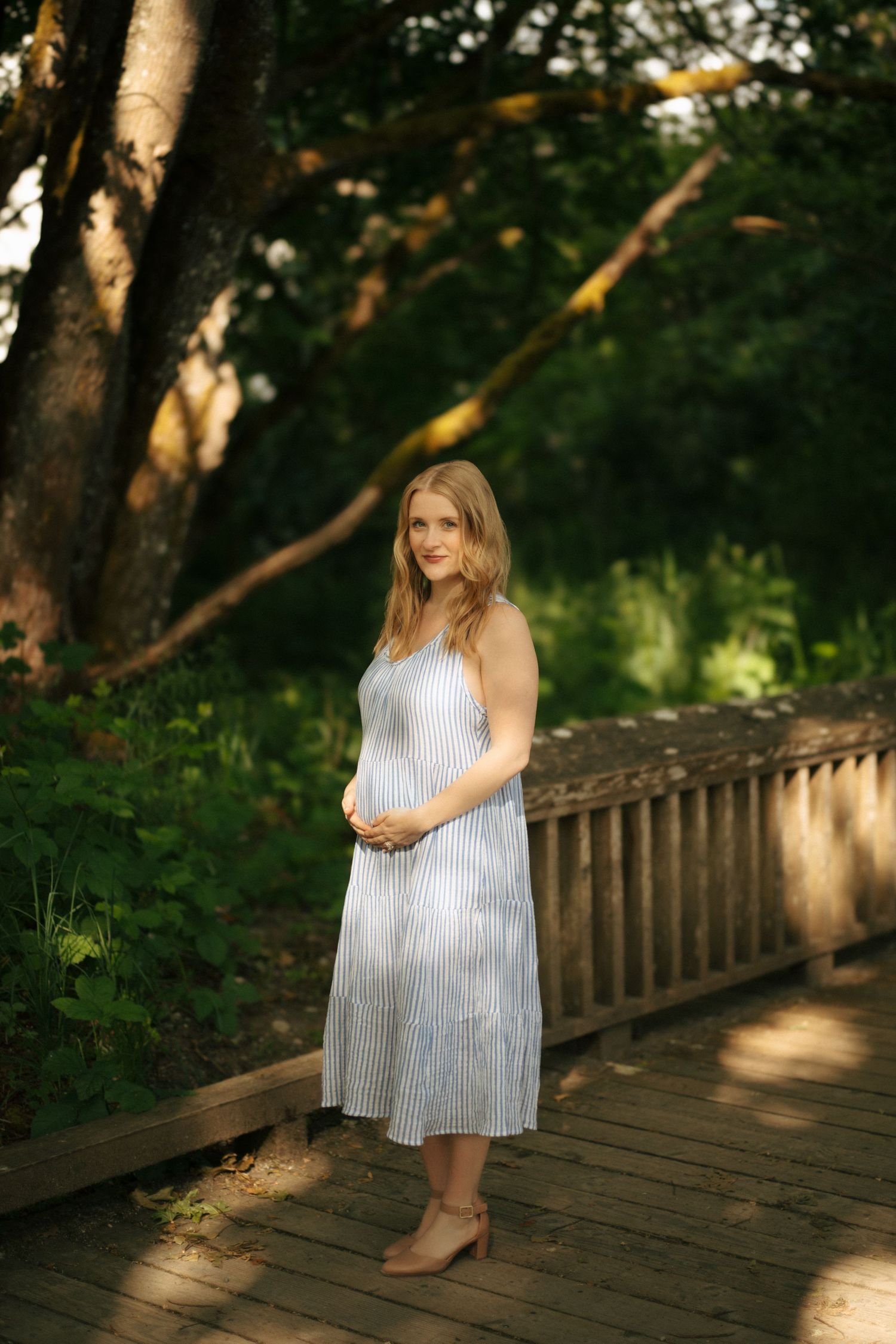 A woman in a striped dress stands in dappled sunlight on a wooden boardwalk surrounded by lush green forest.