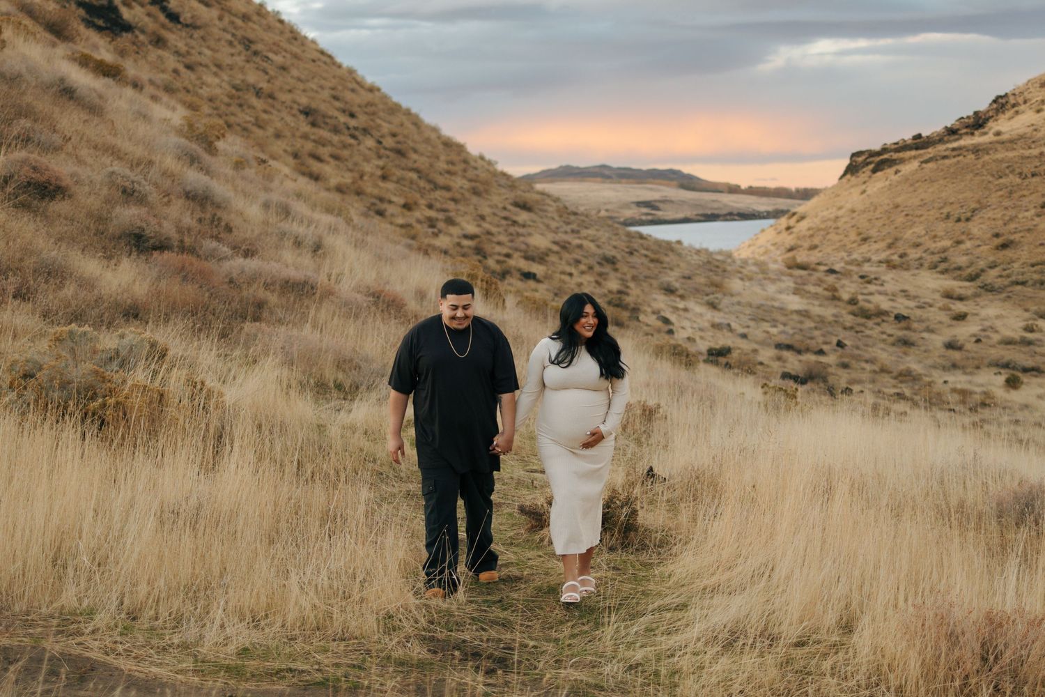 Couple walking through golden grass hills at sunset, pregnant woman in white dress holding belly, scenic river valley backdrop.
