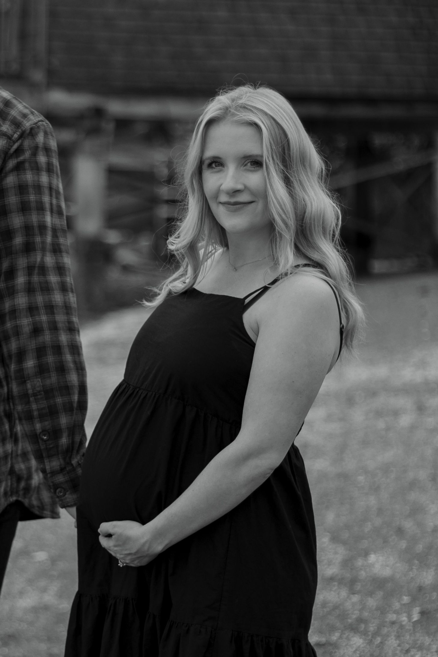 Black and white maternity photo of a smiling blonde woman in a black dress, gently holding her baby bump outdoors.