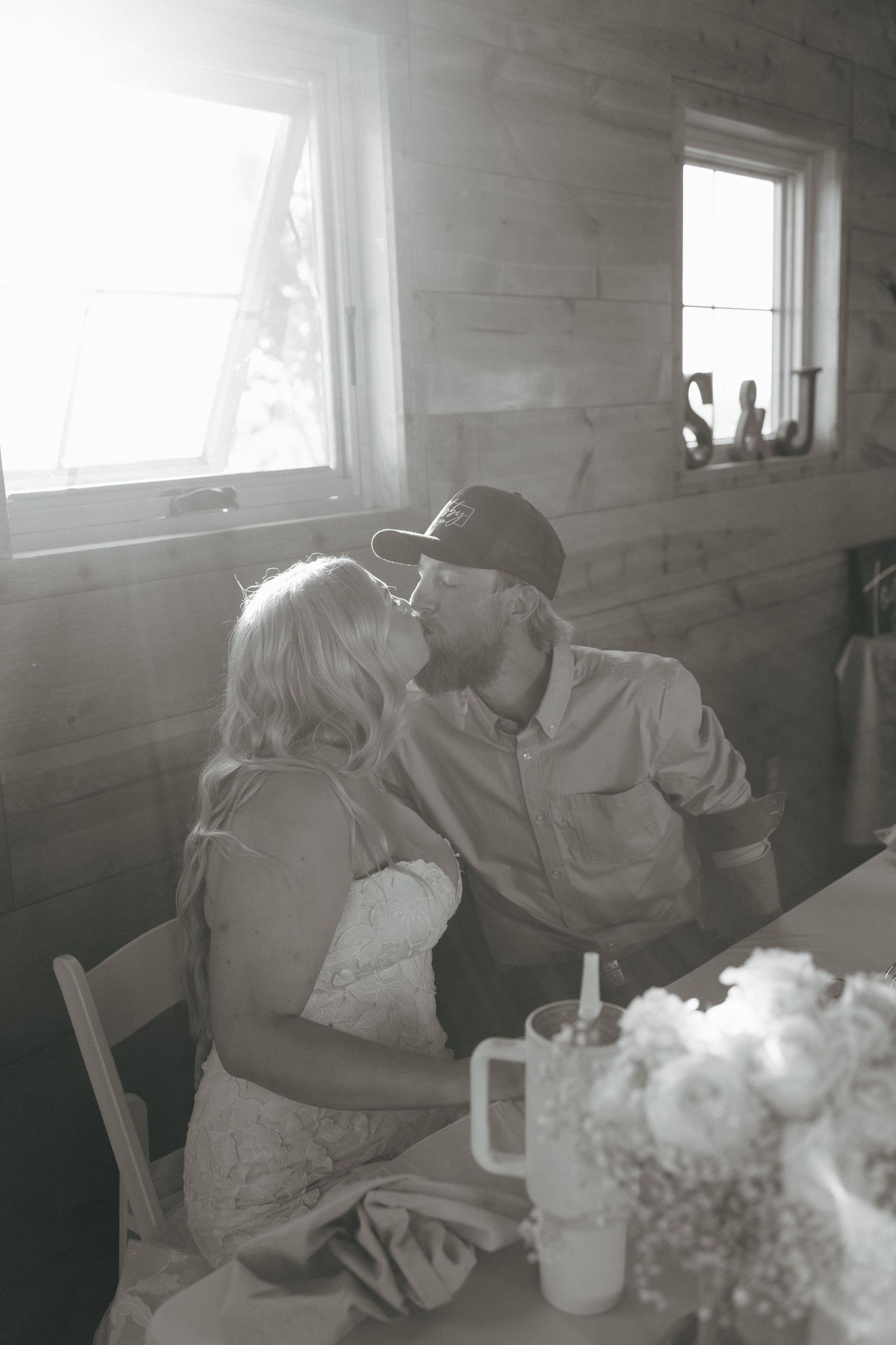 Black and white photo of a couple sharing a kiss at a rustic wooden table with flowers and a mug nearby.