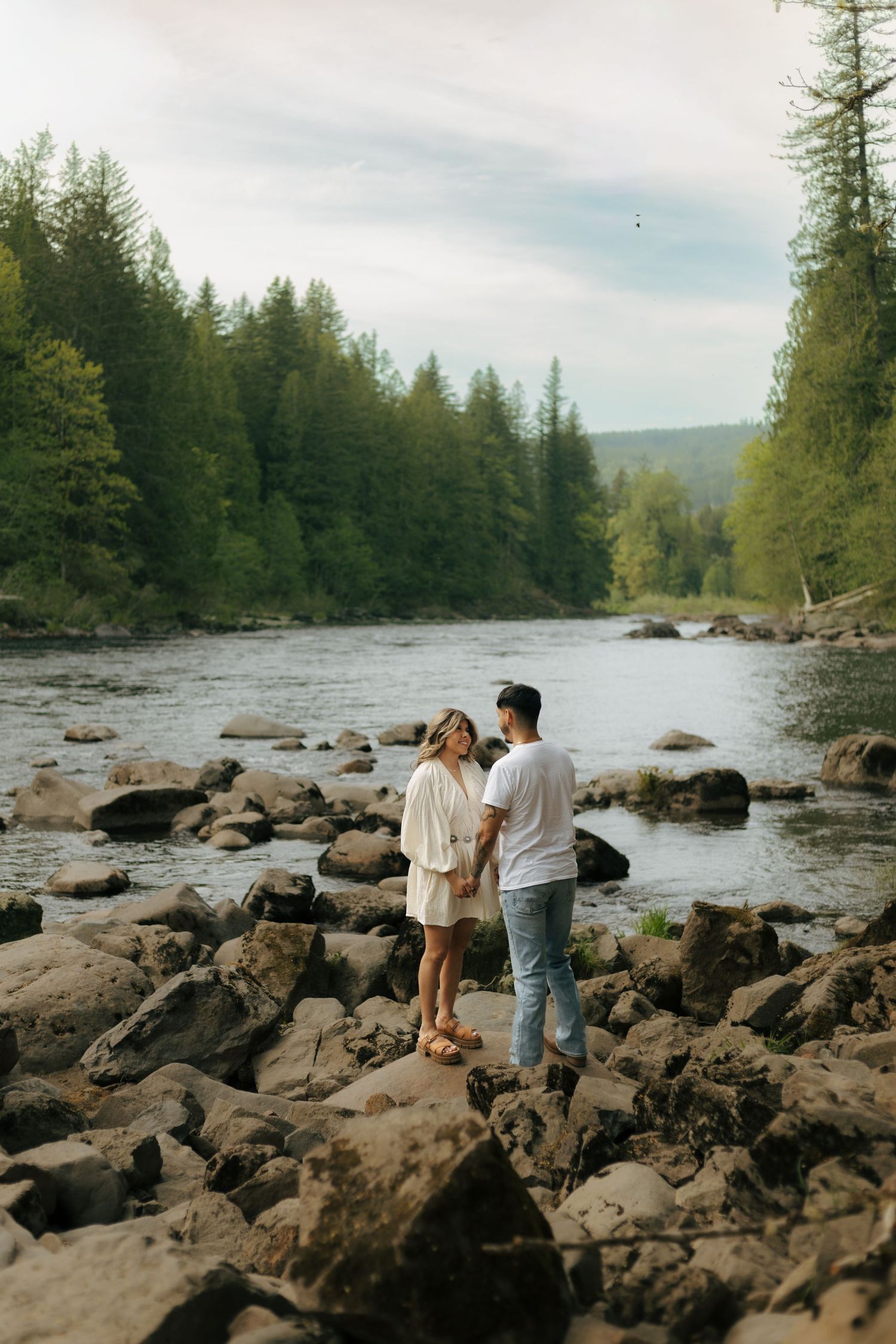 Couple holding hands on rocky riverbank surrounded by lush green forest trees in a scenic outdoor setting.